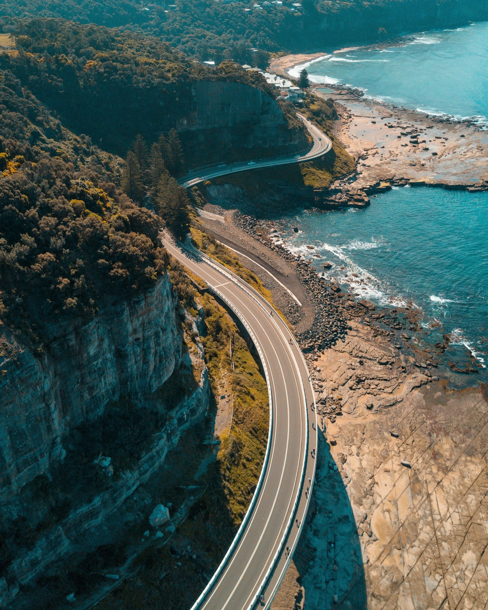Winding coastal highway built into a steep cliffside, overlooking rocky shoreline and the ocean, with trees and buildings in the background.