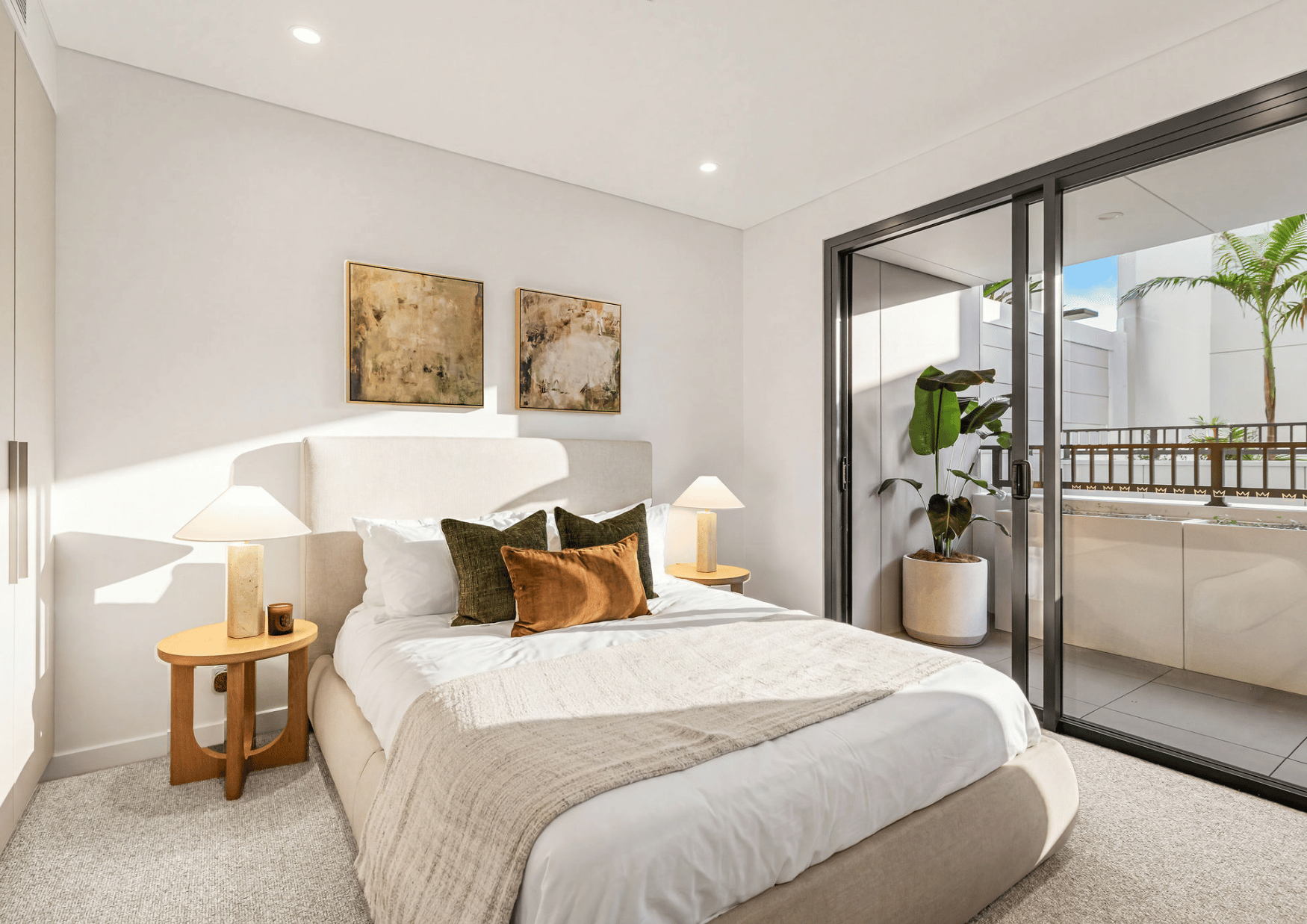 A modern bedroom with a large bed decorated with white and brown pillows, beige blanket, two matching beige nightstands with lamps, abstract art on the white wall, a sliding glass door leading to a balcony with greenery, a potted plant, and bright na
