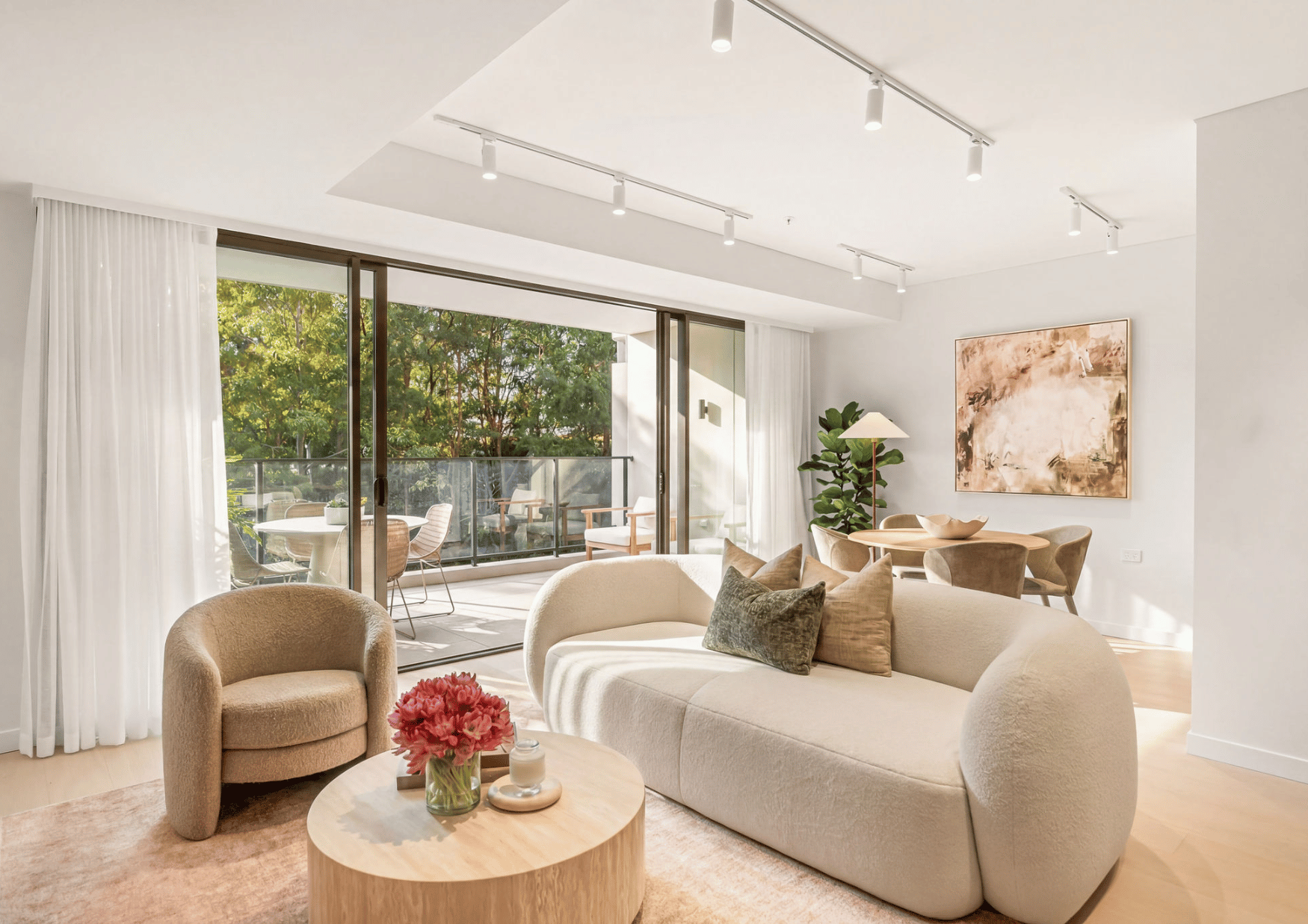 Modern living room with beige sofa, armchair, glass sliding door leading to balcony with outdoor seating, artwork on wall, potted plant, floor lamp, and flowers on coffee table, bright natural lighting