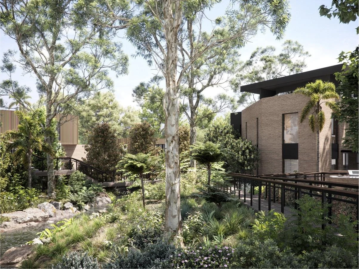 A lush, landscaped outdoor area with tall trees, various green shrubs, and a small stream with rocks. Modern buildings with brick and dark accents are visible in the background, along with a wooden walkway and railing.