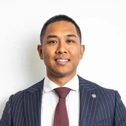 Portrait of a young man in a navy pinstripe suit and red tie, smiling against a white background.