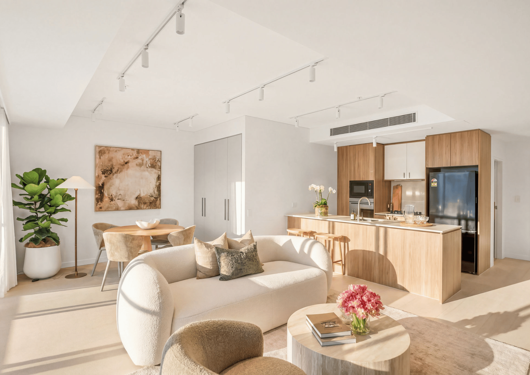 Modern living room and kitchen with white walls, beige furniture, a large green plant, and minimalist decor, illuminated by natural sunlight.