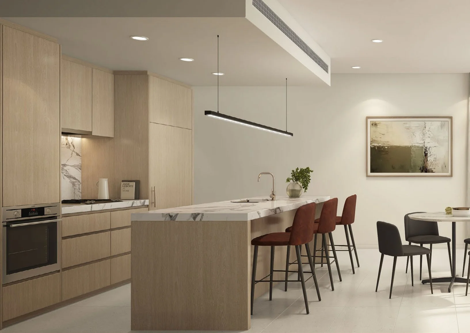 Modern kitchen with light wood cabinets, marble countertops, and a kitchen island with bar stools. There is a sink, a potted plant, and a piece of abstract wall art in the background. Additional dining chairs and a small table are also visible.