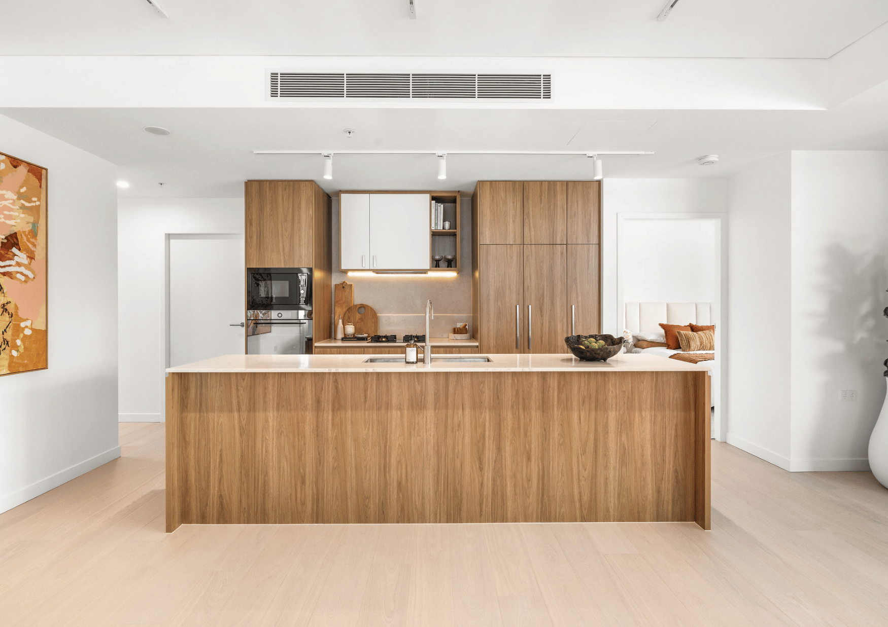 Modern kitchen with wooden cabinets, an island with a beige countertop, and built-in appliances. There is a small seating area with cushions in the background.