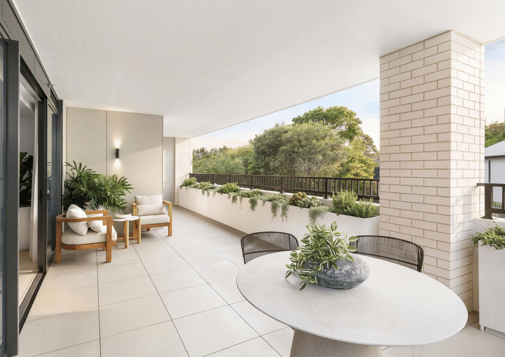 Modern balcony with white tiled floor, white brick columns, black railing, potted plants, and outdoor furniture including a round table with a plant, and cream-colored chairs with wooden arms, overlooking green trees.