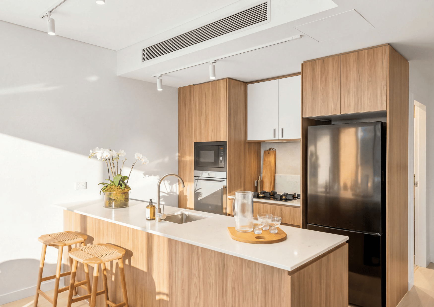 Modern kitchen with white countertops, wooden cabinets, black refrigerator, built-in microwave, gas stove, and small seating area with wicker bar stools. Decor includes a potted orchid and glass pitcher on the counter.