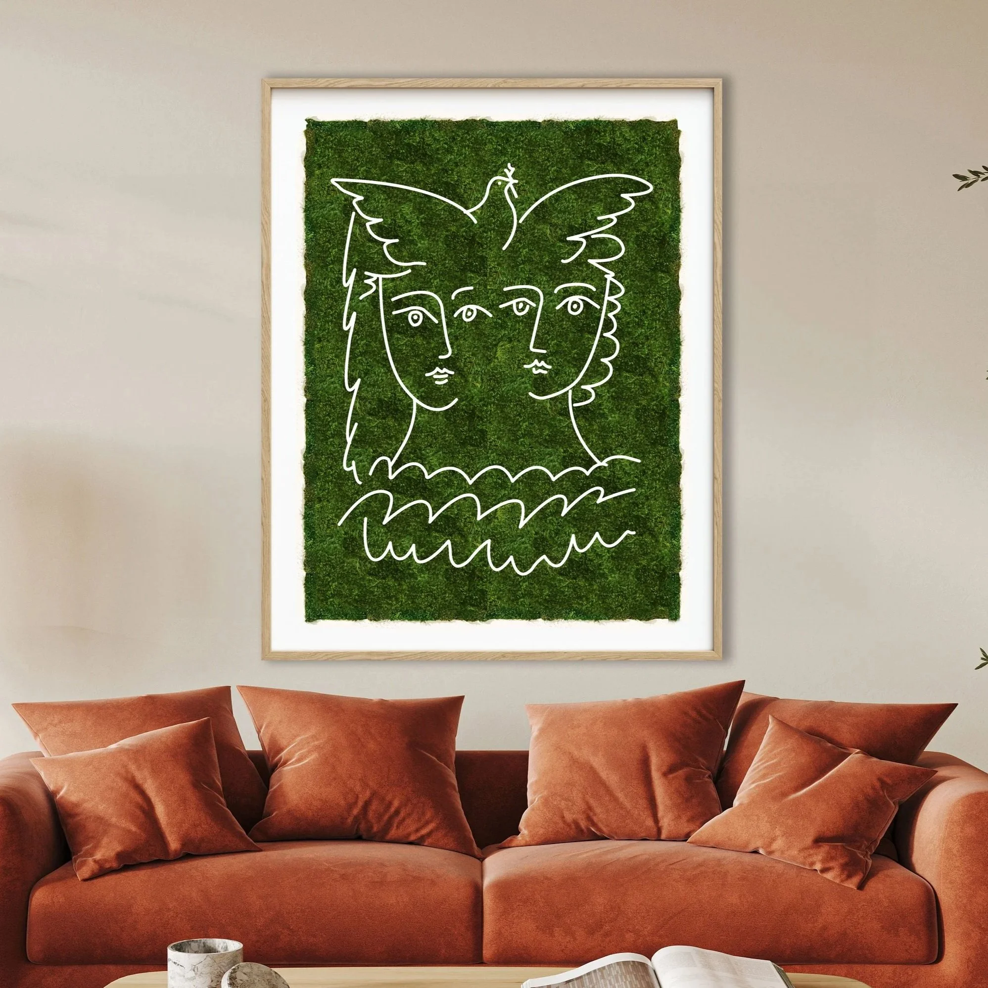 Living room with rust-colored velvet sofa and matching pillows, white coffee table with marble vases and magazine, framed art featuring green moss with line drawing of faces, and a plant on the right side.