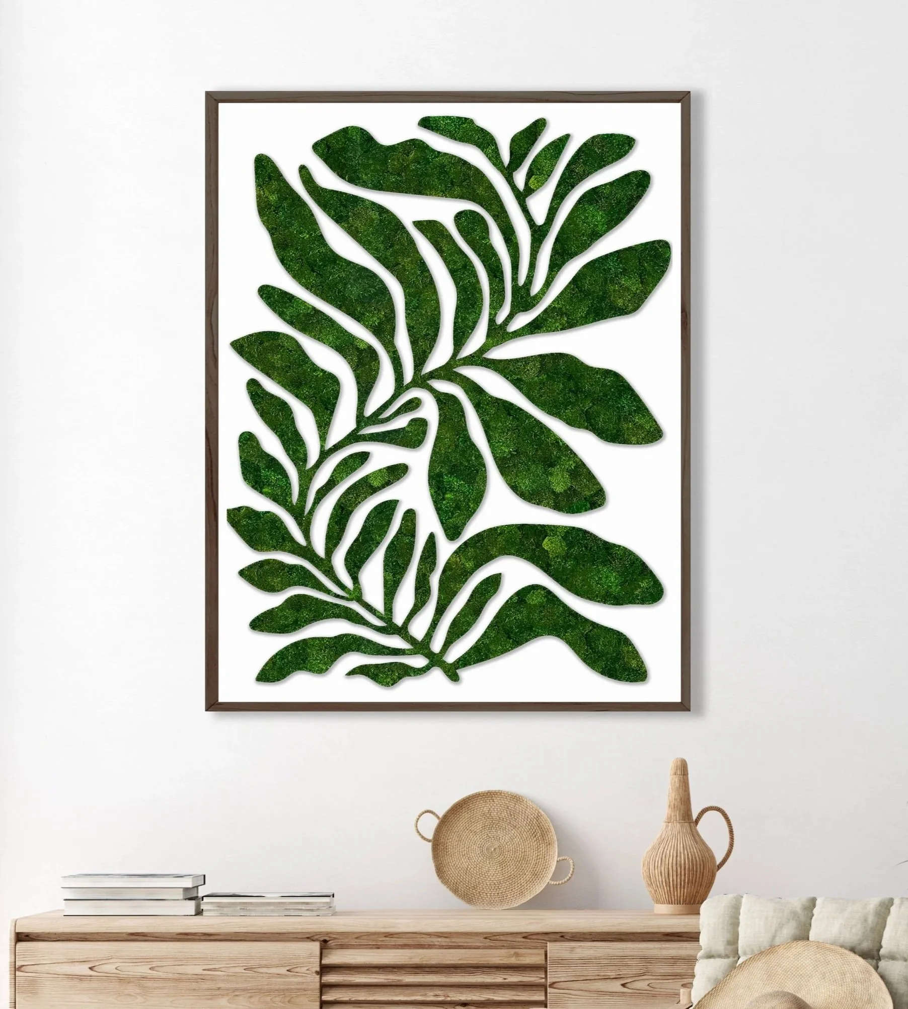 Decorative preserved moss artwork featuring a green botanical pattern on a white background, framed on a white wall above a wooden sideboard with books and wicker vases.