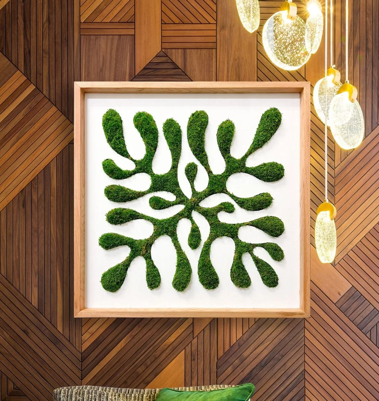 Decorative wall art with green moss arranged in a leaf pattern, framed and mounted on a white background, with warm wooden wall panels and hanging modern light fixtures nearby.