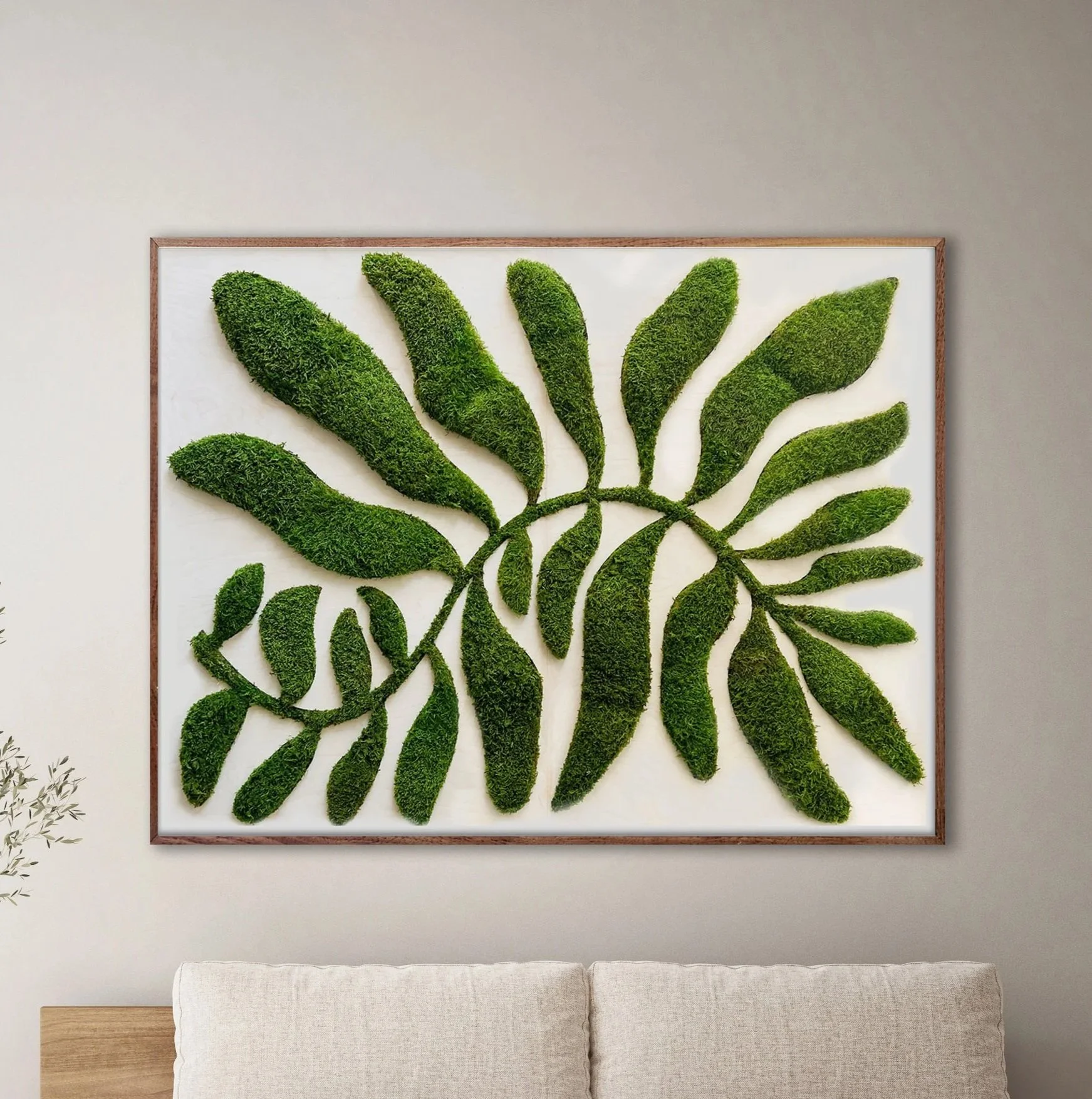 A framed wall art sculpture of preserved green moss arranged as leaves and branches on a white background, hanging on a wall above a beige upholstered bench.