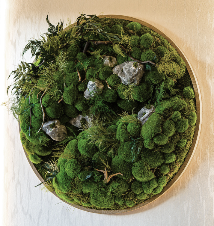 A circular wall decor made of preserved moss, small rocks, and artificial plants, mounted on a wooden frame on a beige wall.