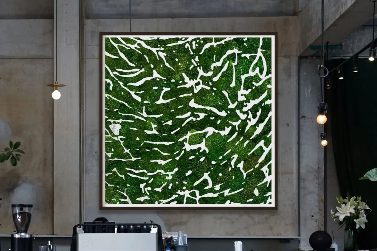 Green and white abstract preserved moss wall art in a modern industrial interior with concrete walls, hanging round light bulbs, and some plants.