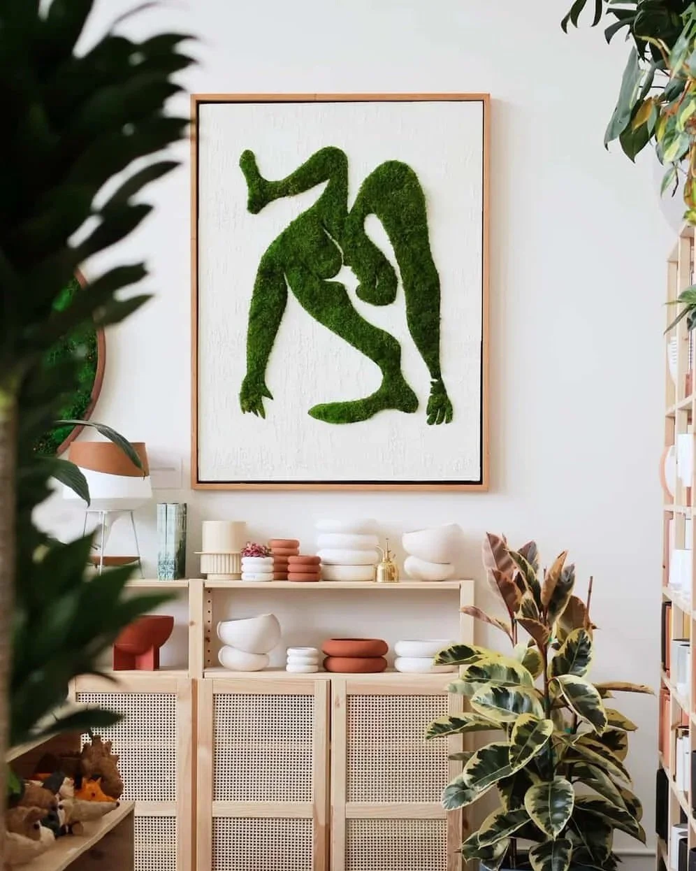 A large framed artwork of a green moss sculpture of two abstract human figures intertwined hangs on a white wall in a room filled with plants and shelves of ceramics.