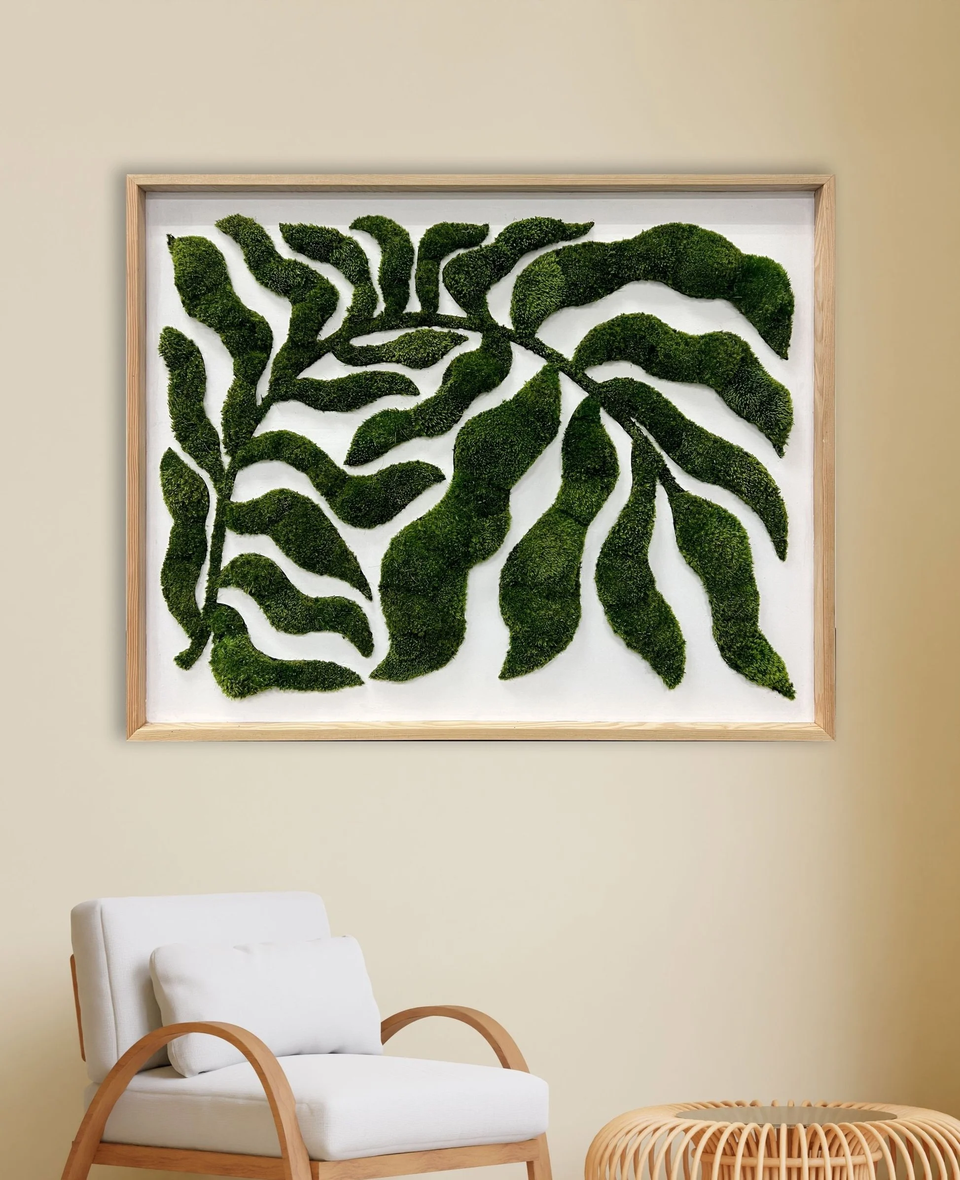 A modern, white cushioned chair with wooden armrests is in the foreground, set against a beige wall. On the wall, there is a framed piece of art featuring moss arranged in the shape of a butterfly. To the right, a small, round, woven rattan table is partially visible.