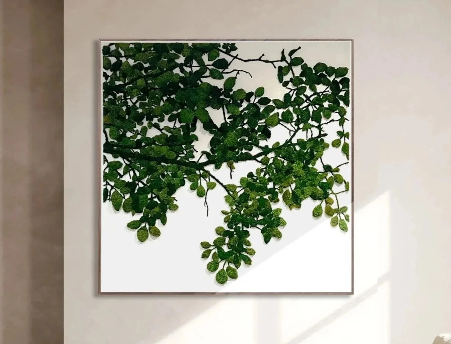 Framed art display of green leaves and branches on a white background, mounted on a beige wall with shadows cast nearby.