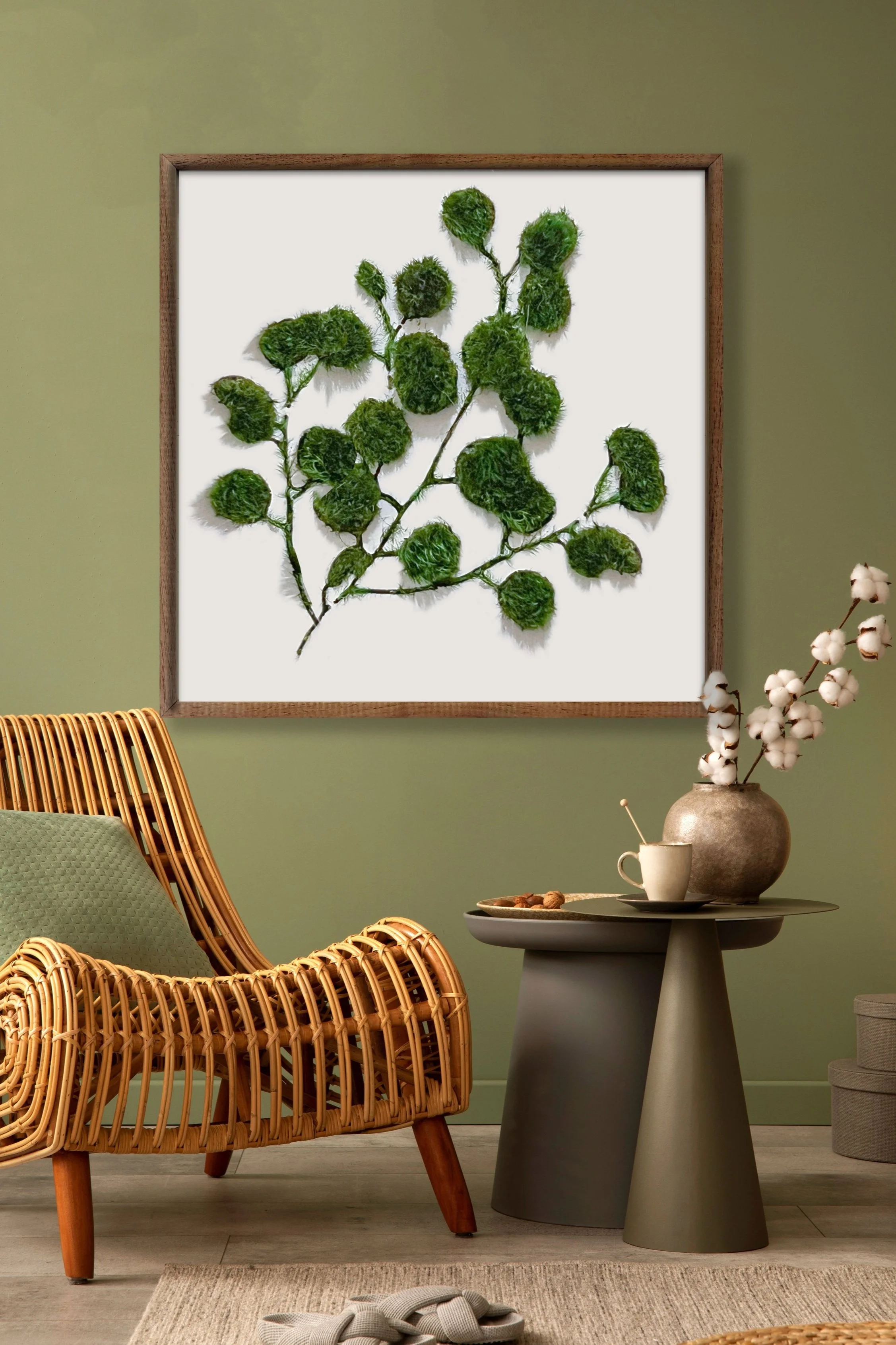 A cozy room with green walls, featuring a framed wall art with preserved moss and twigs, a rattan chair with a green cushion, and a small round table with a beige vase with cotton flowers, a white cup, and a plate of nuts.