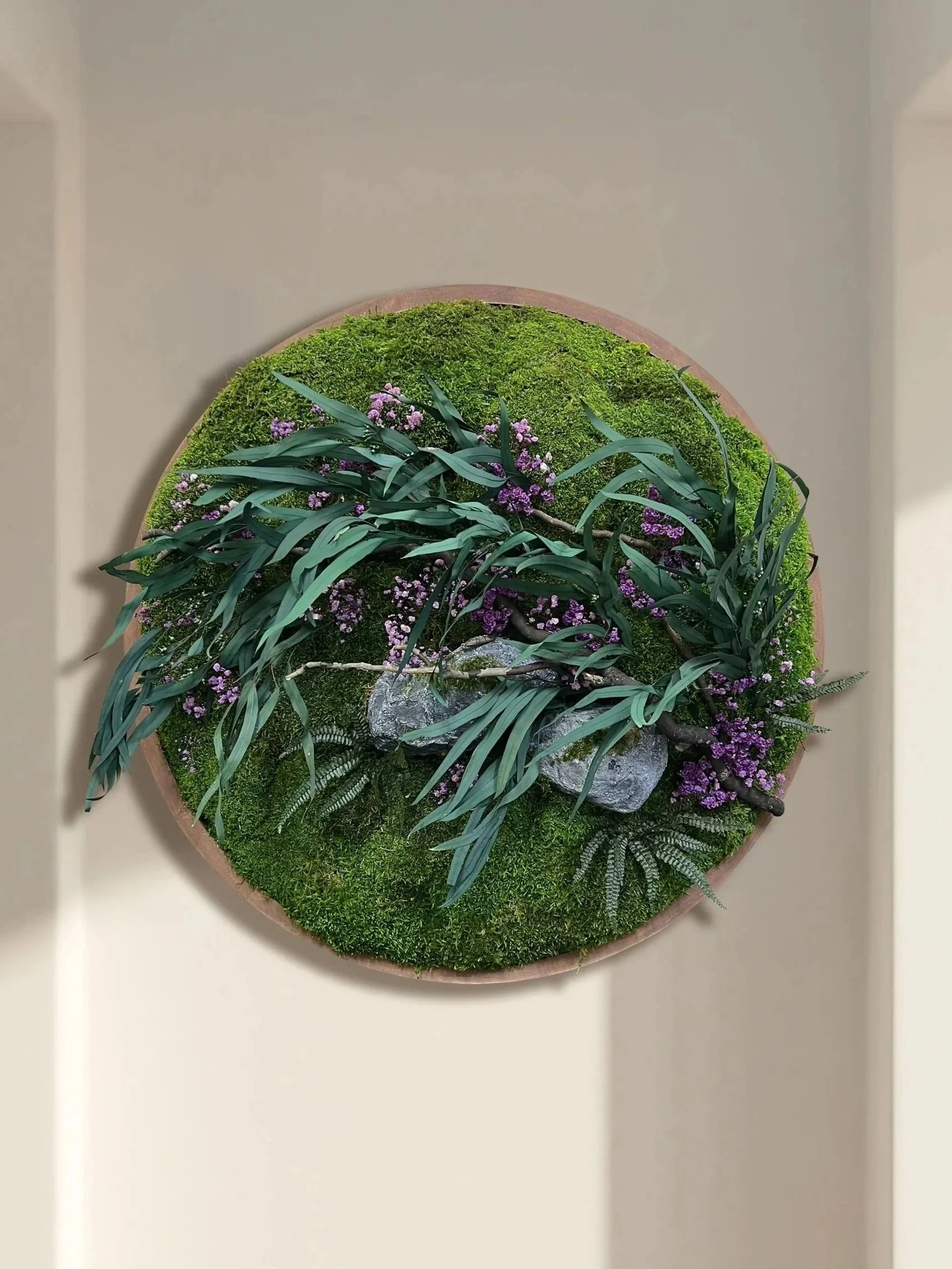 A round display of various mosses, plants, and rocks with purple flowers, arranged decoratively against a beige background.