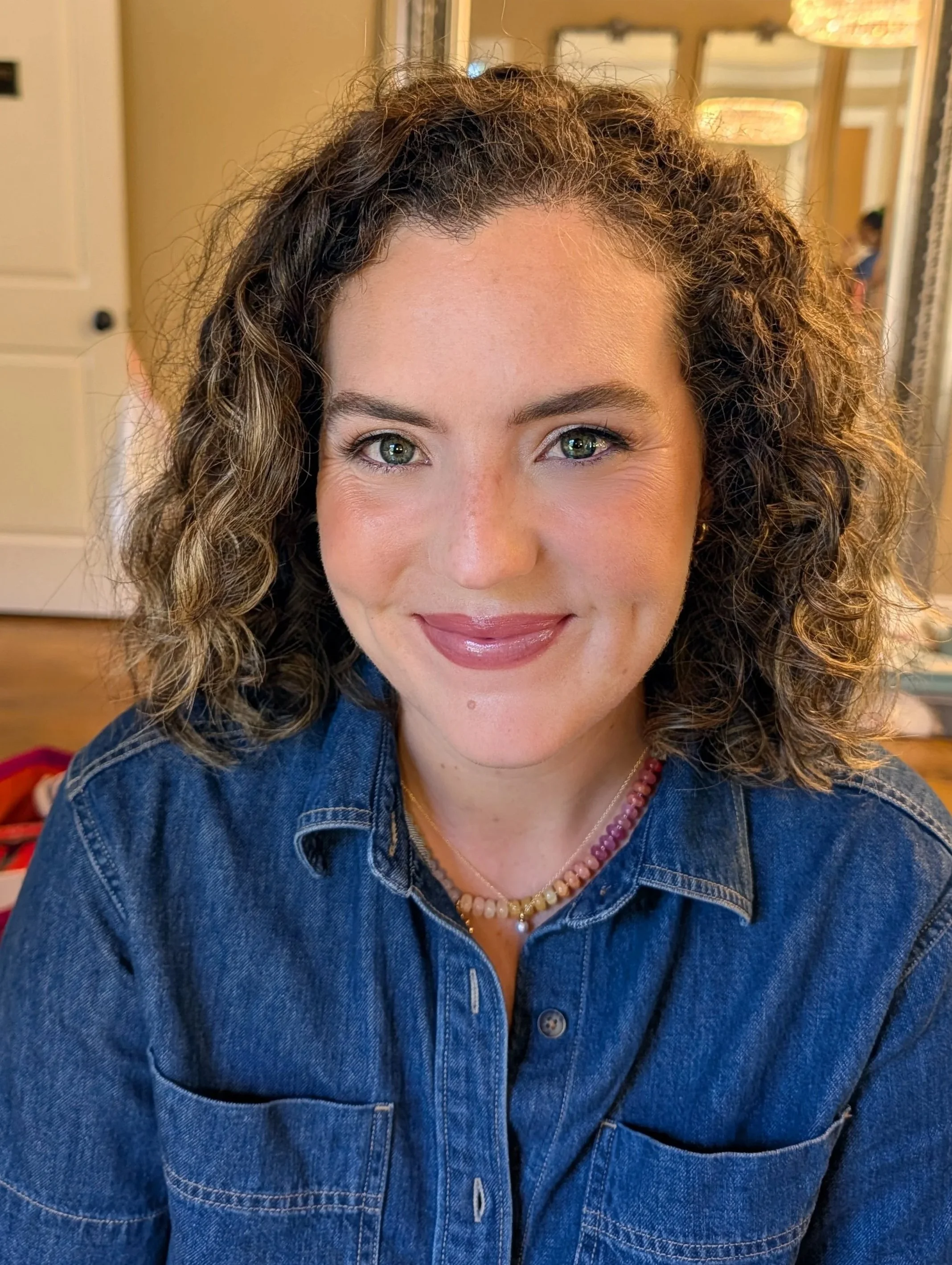 Close-up of a woman with curly brown hair and green eyes, wearing a blue denim jacket and a beaded necklace, smiling at the camera inside a warmly lit room.