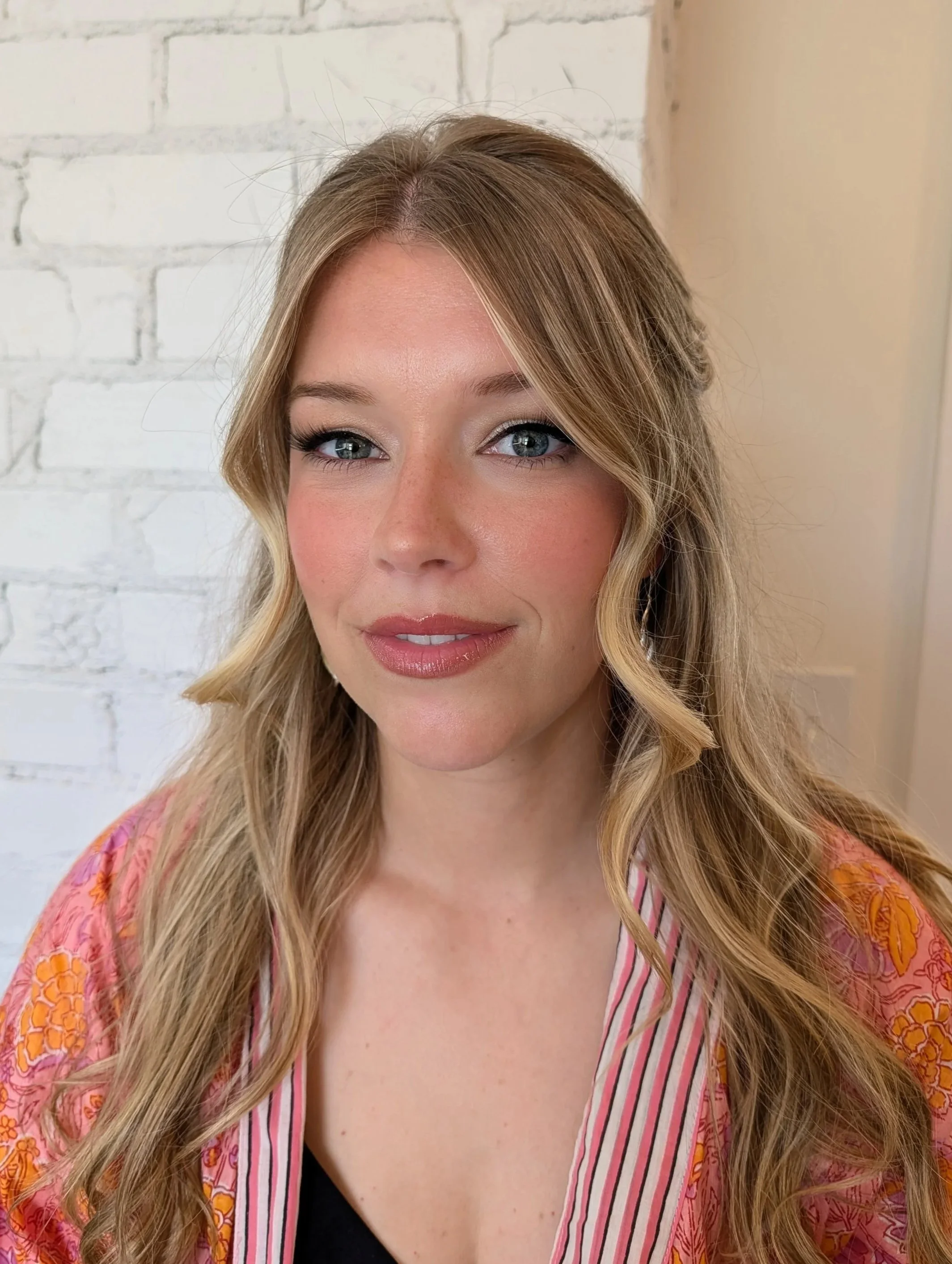 Close-up of a woman with long, wavy blonde hair, wearing makeup, a pink patterned top with vertical striped accents, and a black top underneath, standing against a white brick wall.