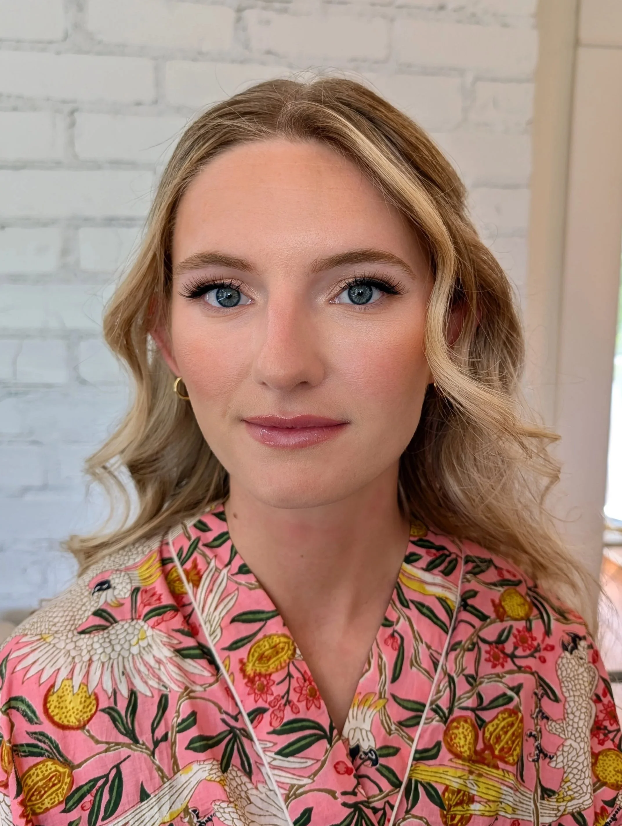 A woman with blonde hair, blue eyes, wearing makeup including eyeliner and lipstick, and a pink blouse with a floral and bird pattern, standing against a white brick wall.