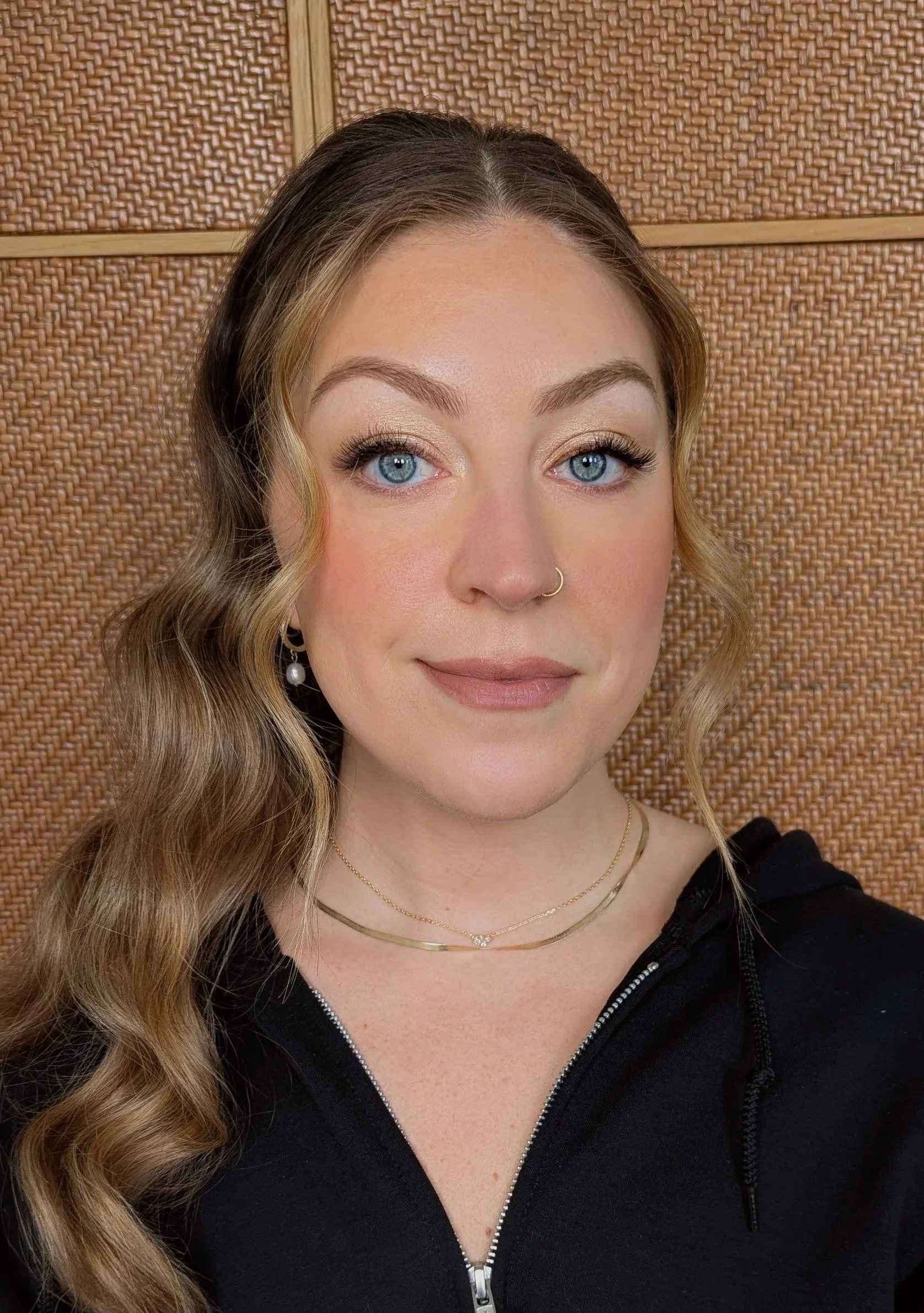 A young woman with blue eyes and long wavy hair poses for a selfie against a woven wooden wall. She wears makeup, a gold necklace, and a black zip-up hoodie.
