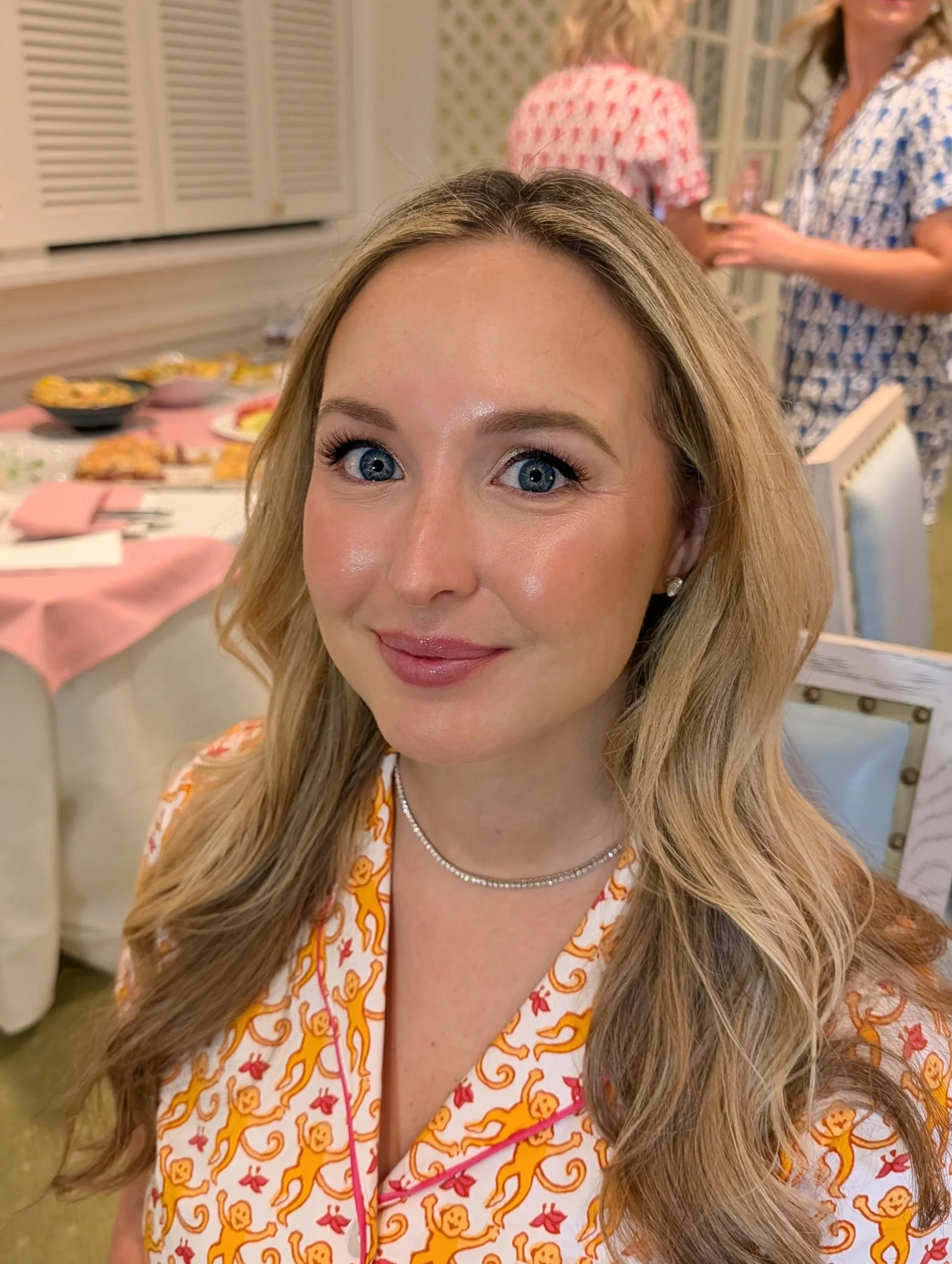 A woman with long blonde hair and blue eyes, smiling, wearing a white dress with orange lion and red floral patterns, in a room with a pink tablecloth, food, and two women in patterned dresses in the background.