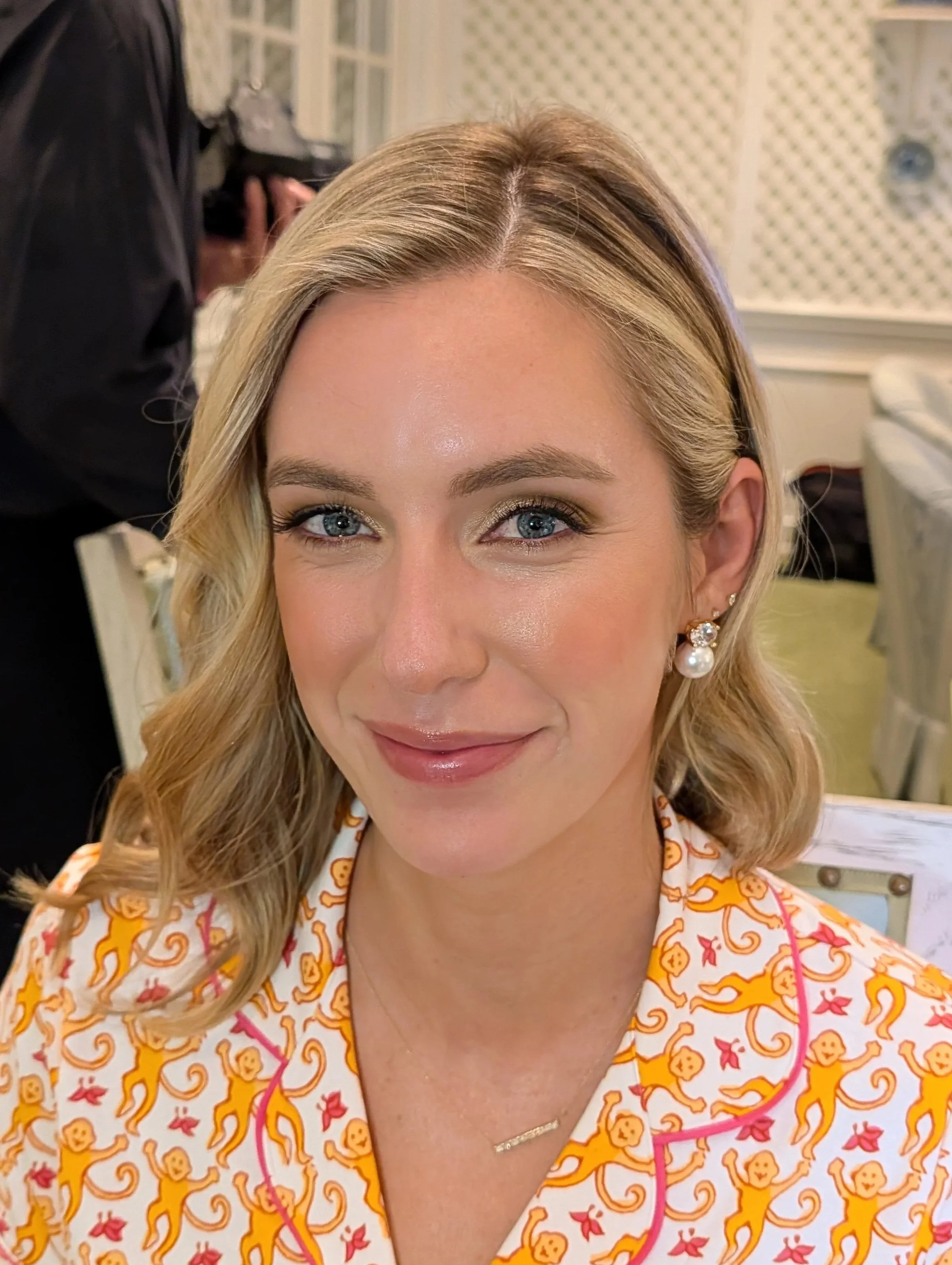 Close-up of a woman with blonde hair and blue eyes, smiling, wearing festive pajamas with a gingerbread men pattern, earrings, and a necklace, in a well-lit room with a patterned wall and a woman in black in the background.