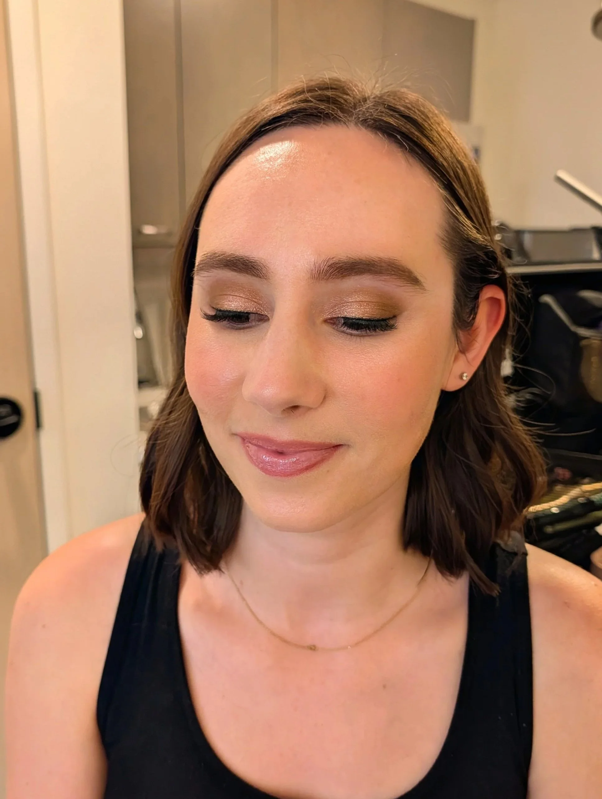 A woman with shoulder-length brown hair and makeup, wearing a black top and a delicate necklace, smiling softly with her eyes closed, in an indoor setting.