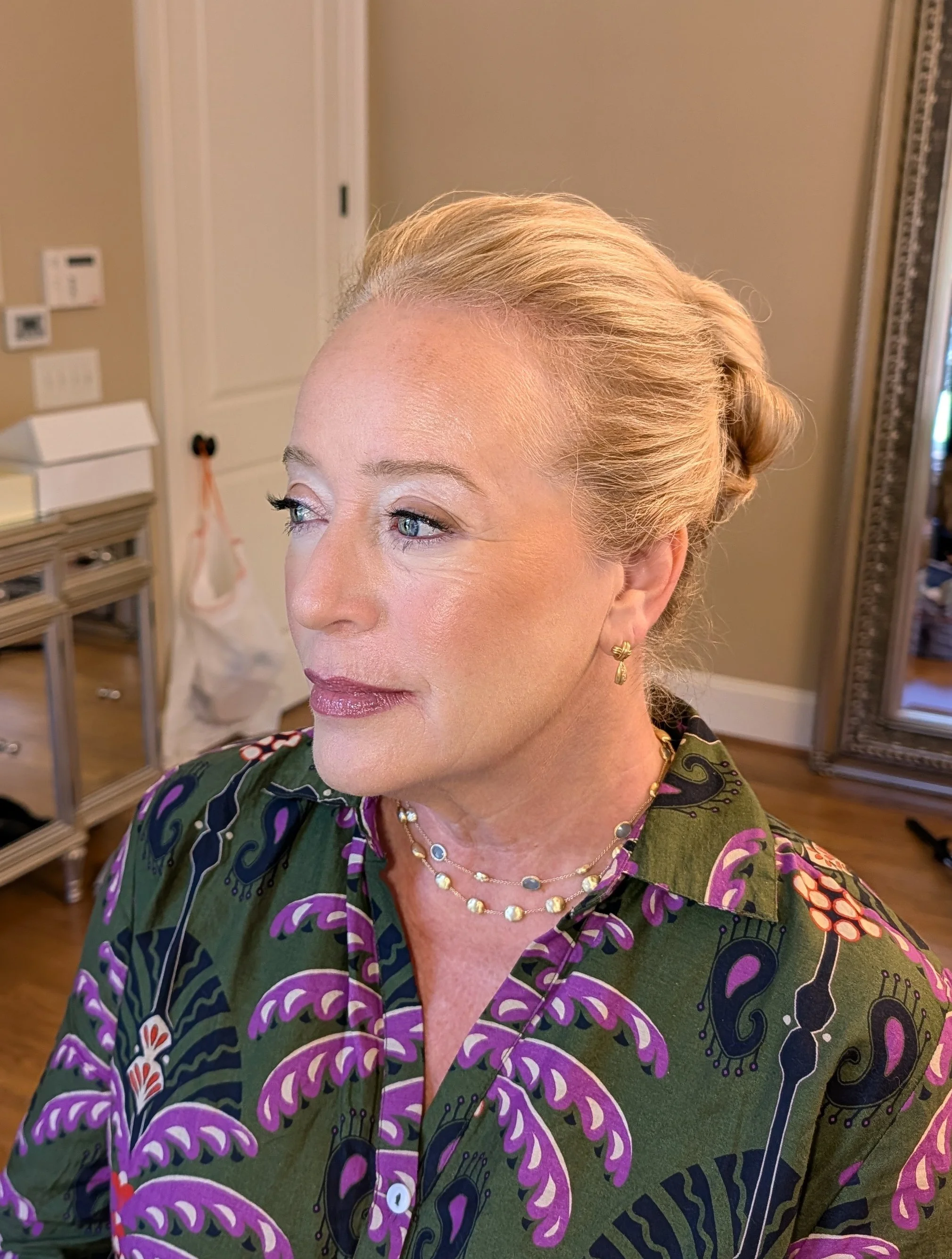 A woman with blonde hair styled in a vintage rolled updo, wearing gold earrings and layered necklaces, dressed in a colorful green and purple patterned blouse, standing indoors with a mirror and wooden furniture in the background.