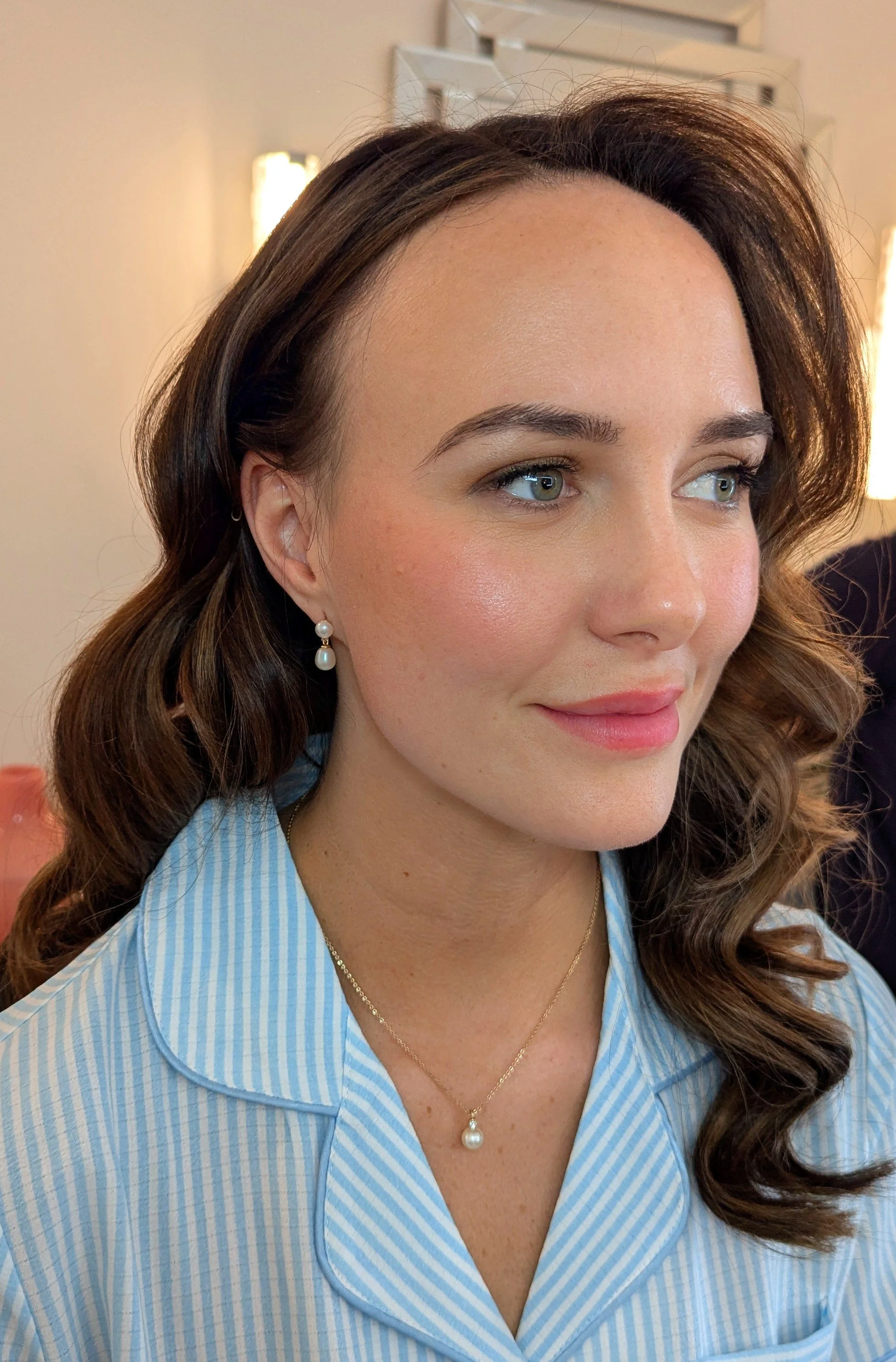 Close-up of a woman with long wavy brown hair, wearing pearl earrings, a gold necklace, and a light blue striped shirt, smiling softly.