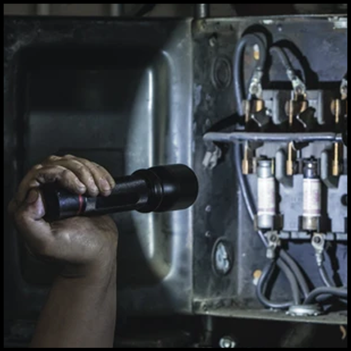 Hand holding flashlight illuminating electrical panel with wires and fuses.