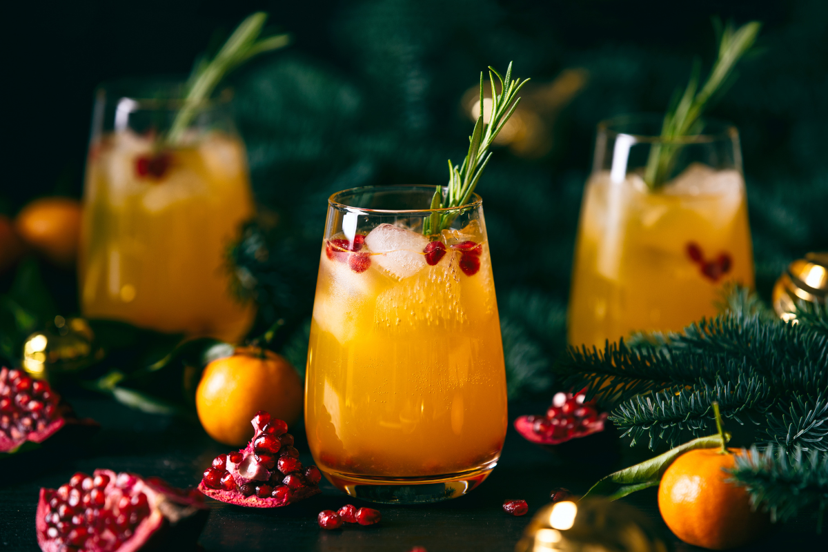 Festive orange cocktail with ice, pomegranate seeds, and rosemary garnish, surrounded by pomegranate halves and tangerines on a dark background with greenery.