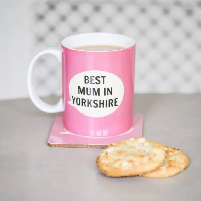 Pink mug with "Best Mum in Yorkshire" text, placed on coaster with two cookies nearby.