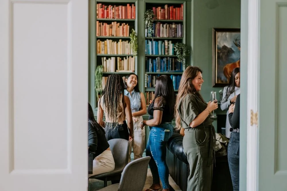 Sara Maxwell in green jumpsuit with a drink in a library mingling with other women