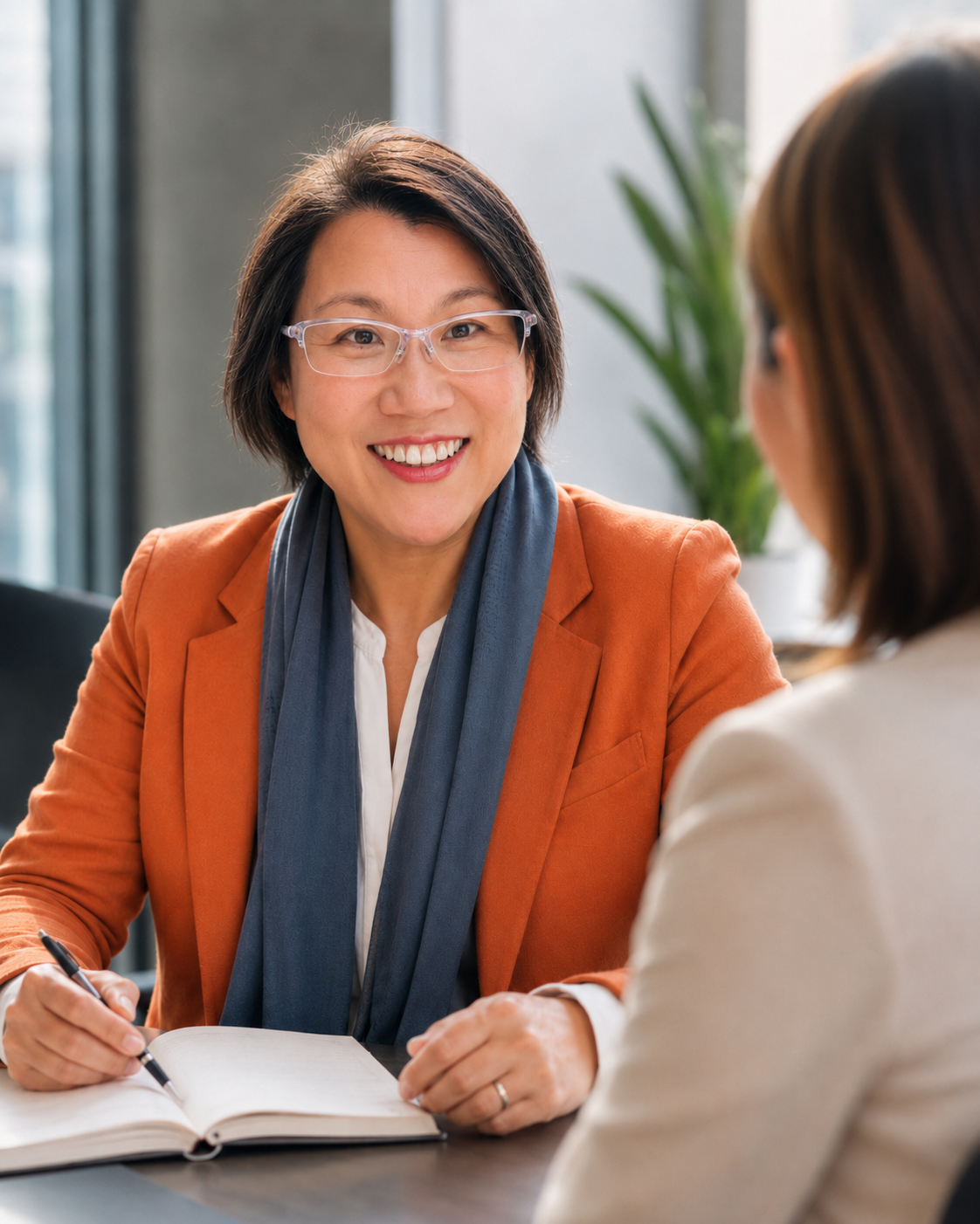 A woman with short dark hair, glasses, and a bright orange blazer smiling during a conversation with another woman in an office setting.