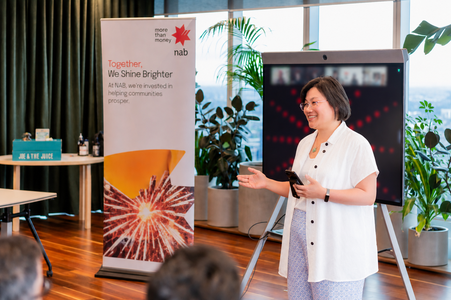 A woman giving a presentation in an office or conference room, standing next to a large digital screen and a banner with the NAB logo and slogan.