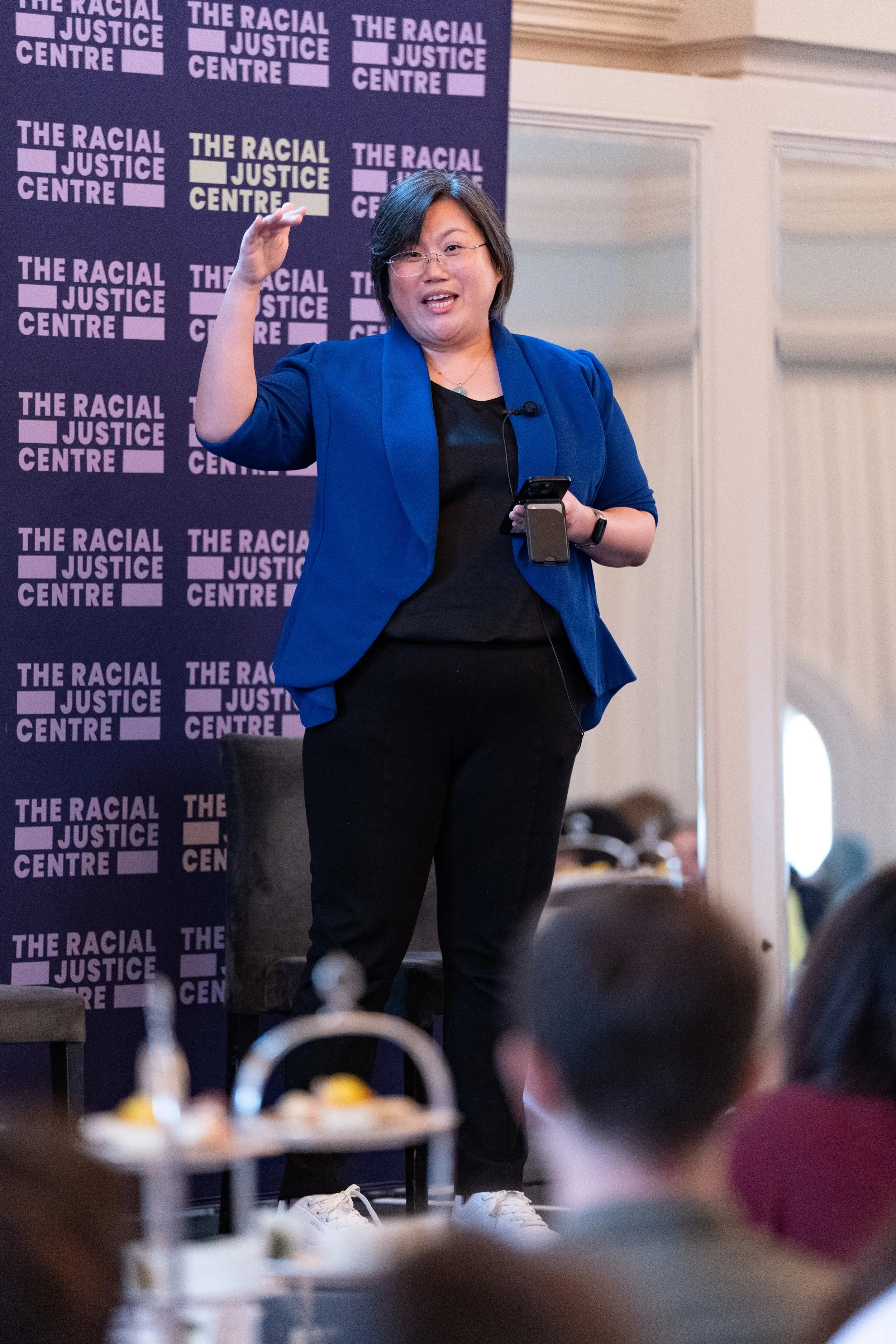 A woman speaking at an event with a backdrop that reads 'The Racial Justice Centre.' She is wearing glasses, a blue blazer, black pants, and white sneakers, and is holding a microphone and a smartphone. In the foreground, there are blurred plates of food.