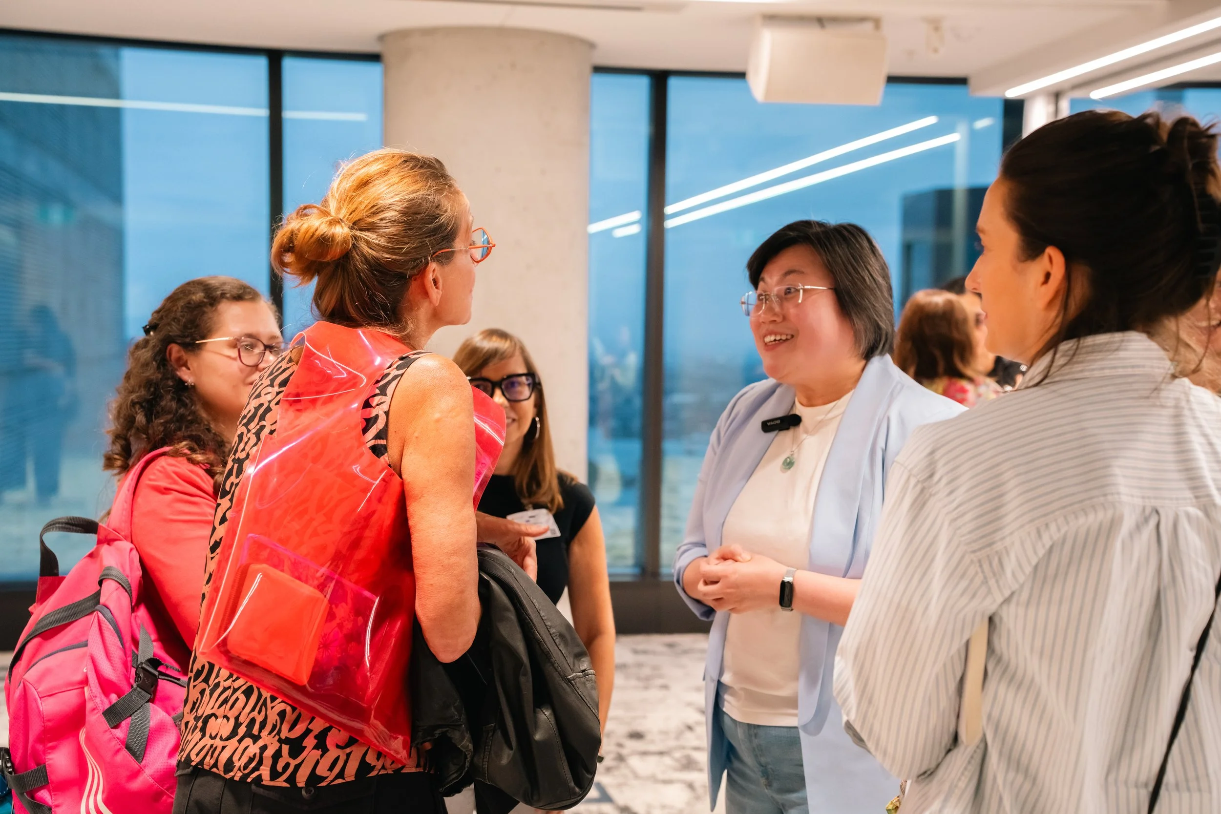 Group of women talking and smiling in an indoor setting with large windows, some carrying backpacks, one woman gesturing with her hands, and others listening attentively.