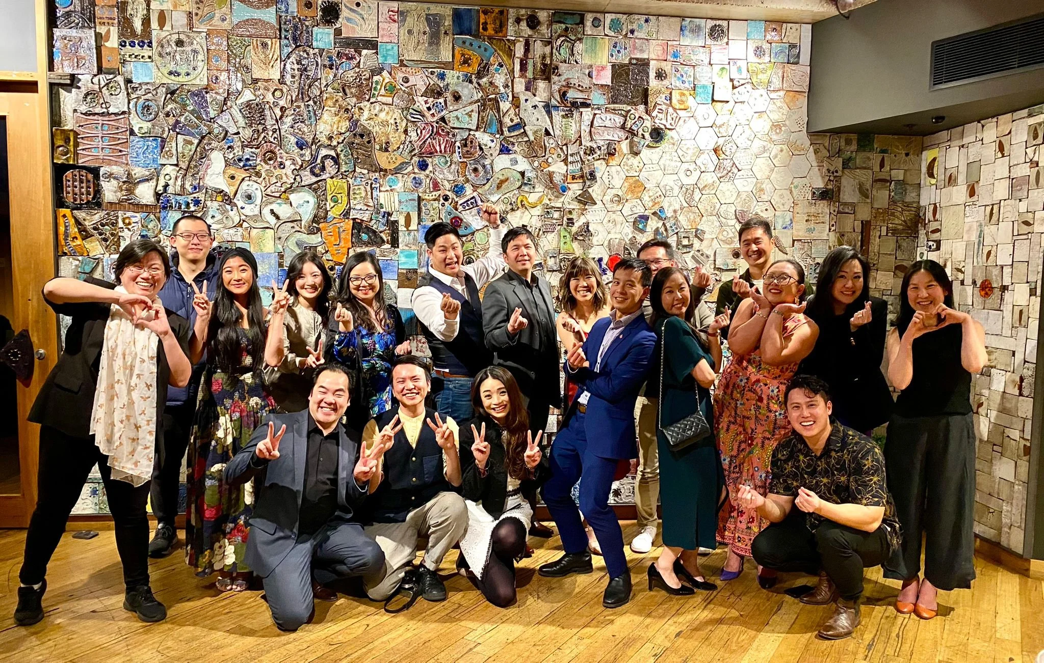A group of people posing together in front of a mosaic wall inside a building. The group is smiling and making various hand gestures, including peace signs and heart shapes.