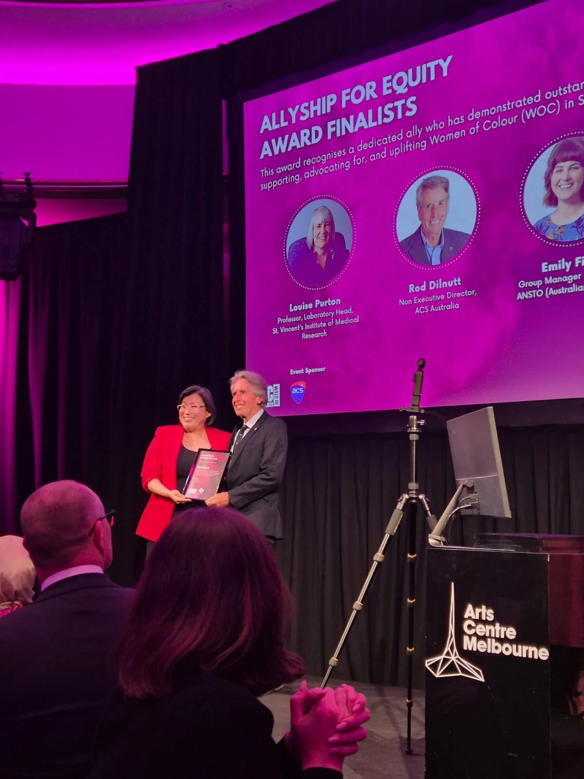 A woman in a red blazer and a man in a dark suit are standing on stage at the Arts Centre Melbourne, holding an award. Behind them, there is a large screen displaying information about the Allyship for Equity Award finalists, with headshots and names of the finalists. Audience members are seated in front of the stage, and there are lighting and curtain decorations in the background.