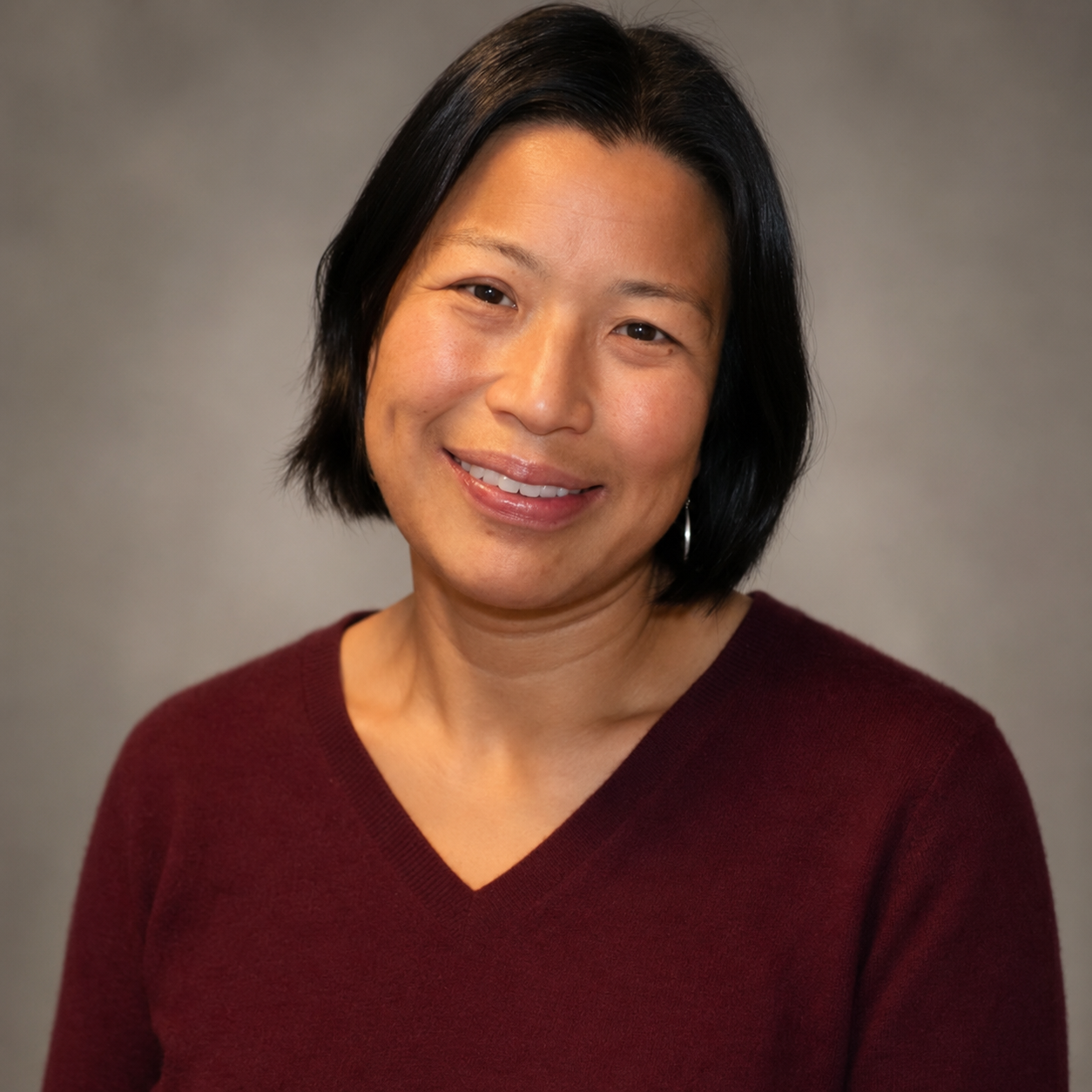 Portrait of a woman with short black hair, wearing a maroon V-neck sweater, smiling, against a gray background.