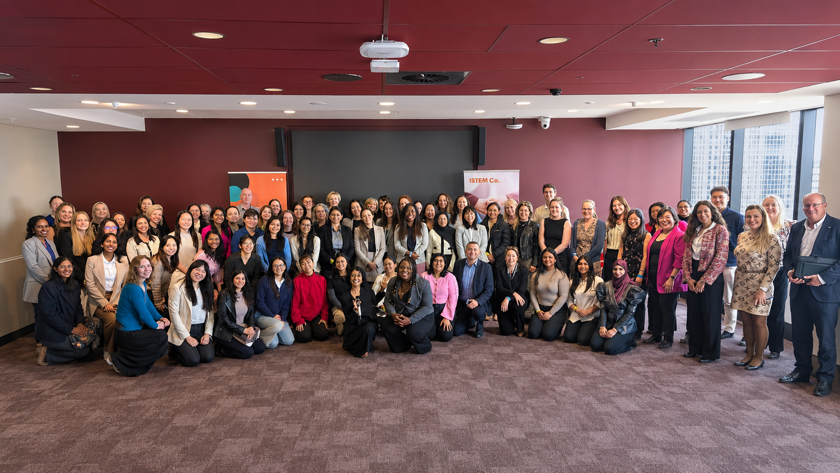 A large group of professional women and men posing for a photo in a conference room with large windows and a city view. Some people are seated on the floor in front, while others stand behind. The room has a maroon accent wall, a large screen, and banners that say 'ISTEM Co.' and feature colorful designs.
