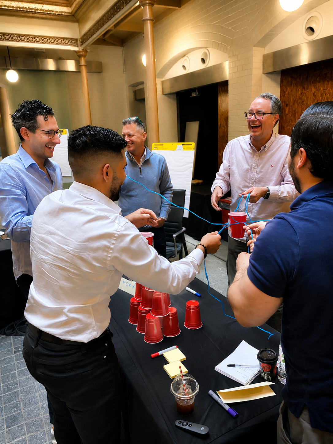 A group of people smiling and having fun playing a team-building game with red plastic cups stacked on a table, connected with blue strings in an indoor setting.