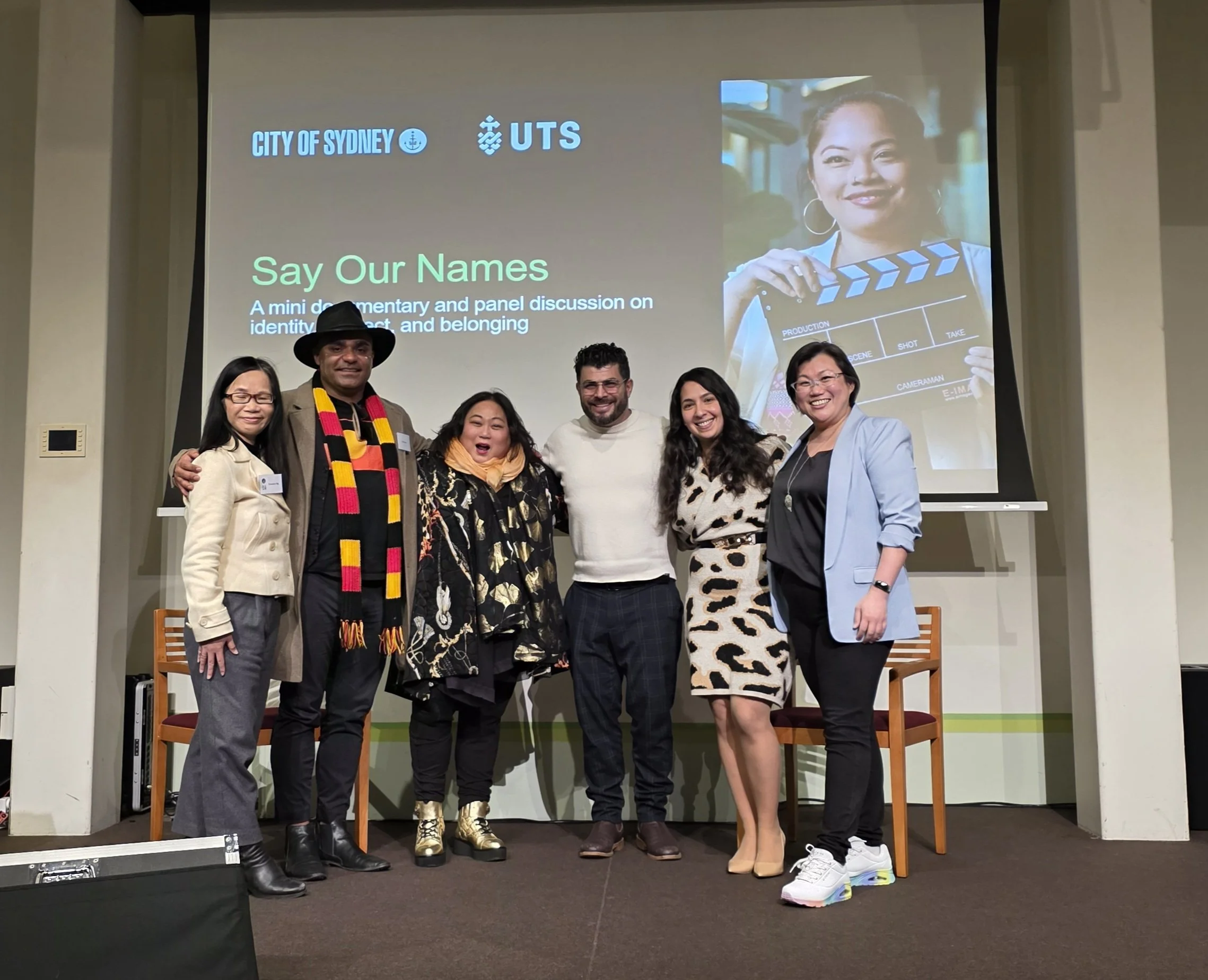 A group of six diverse people standing side by side, smiling, in front of a presentation screen that reads 'Say Our Names' and features logos of the City of Sydney and UTS. The screen also has images of a woman holding a clapboard and people sitting in a room.