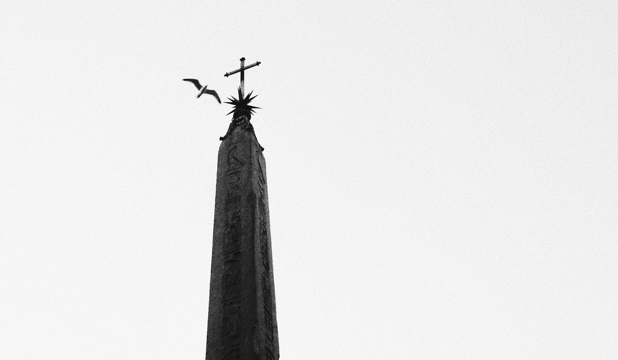 Black-and-white photo of an obelisk with a cross on top, birds flying nearby against the sky.