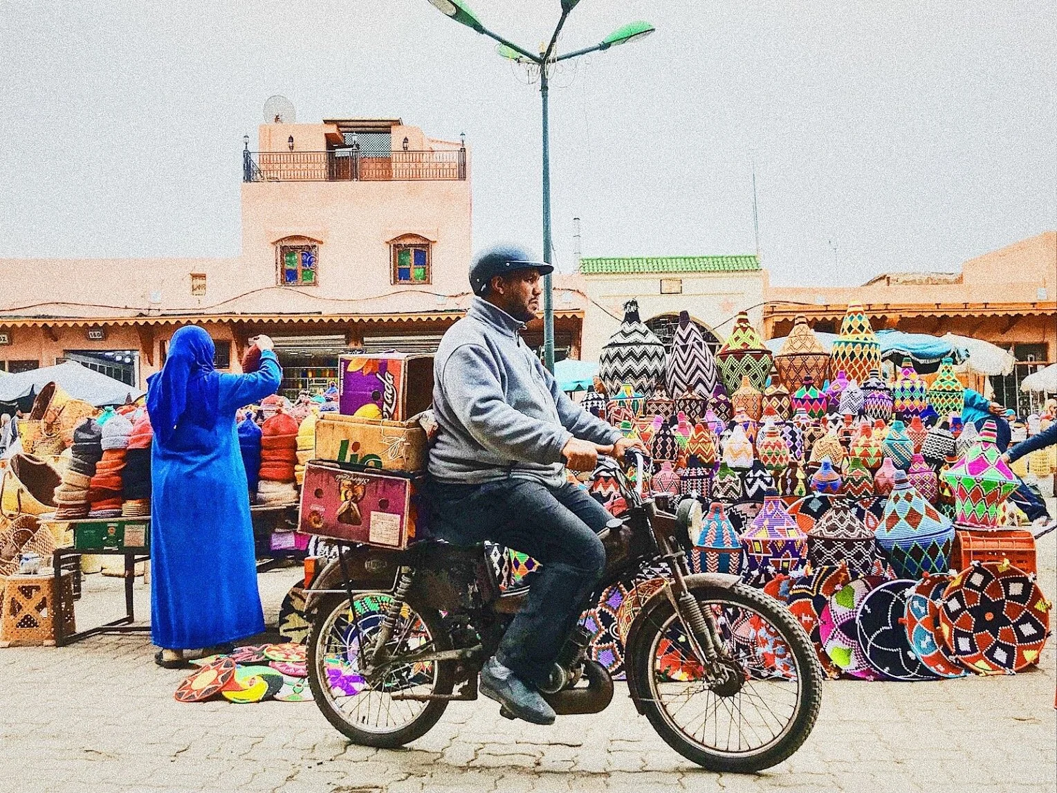 Man on a motorcycle passing a Moroccan market stall with colorful woven baskets and traditional pottery.