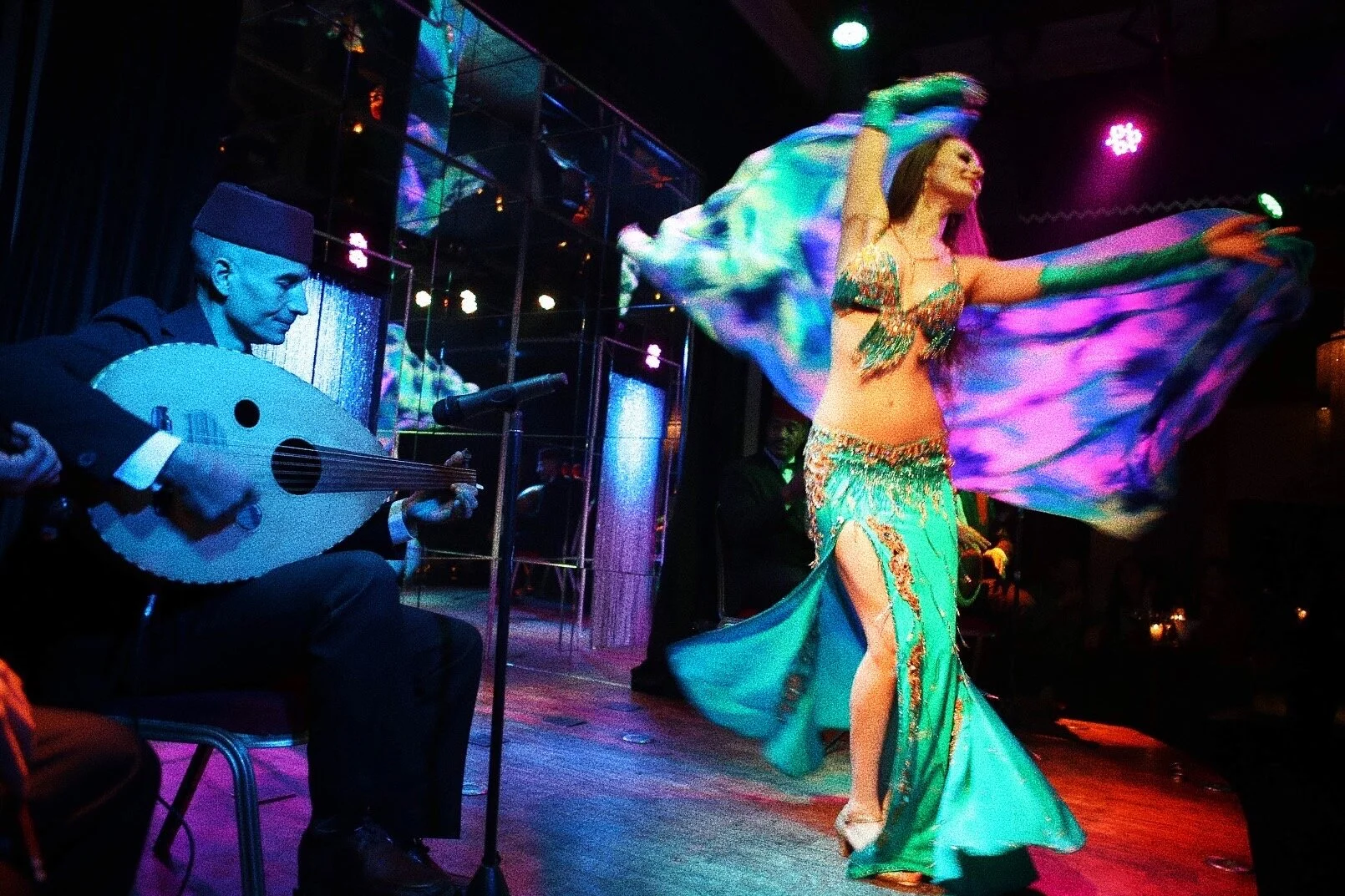 A musician playing an oud sits on stage next to a belly dancer wearing a vibrant costume with a flowing veil. The scene is set in a dimly lit performance venue with colorful lighting.