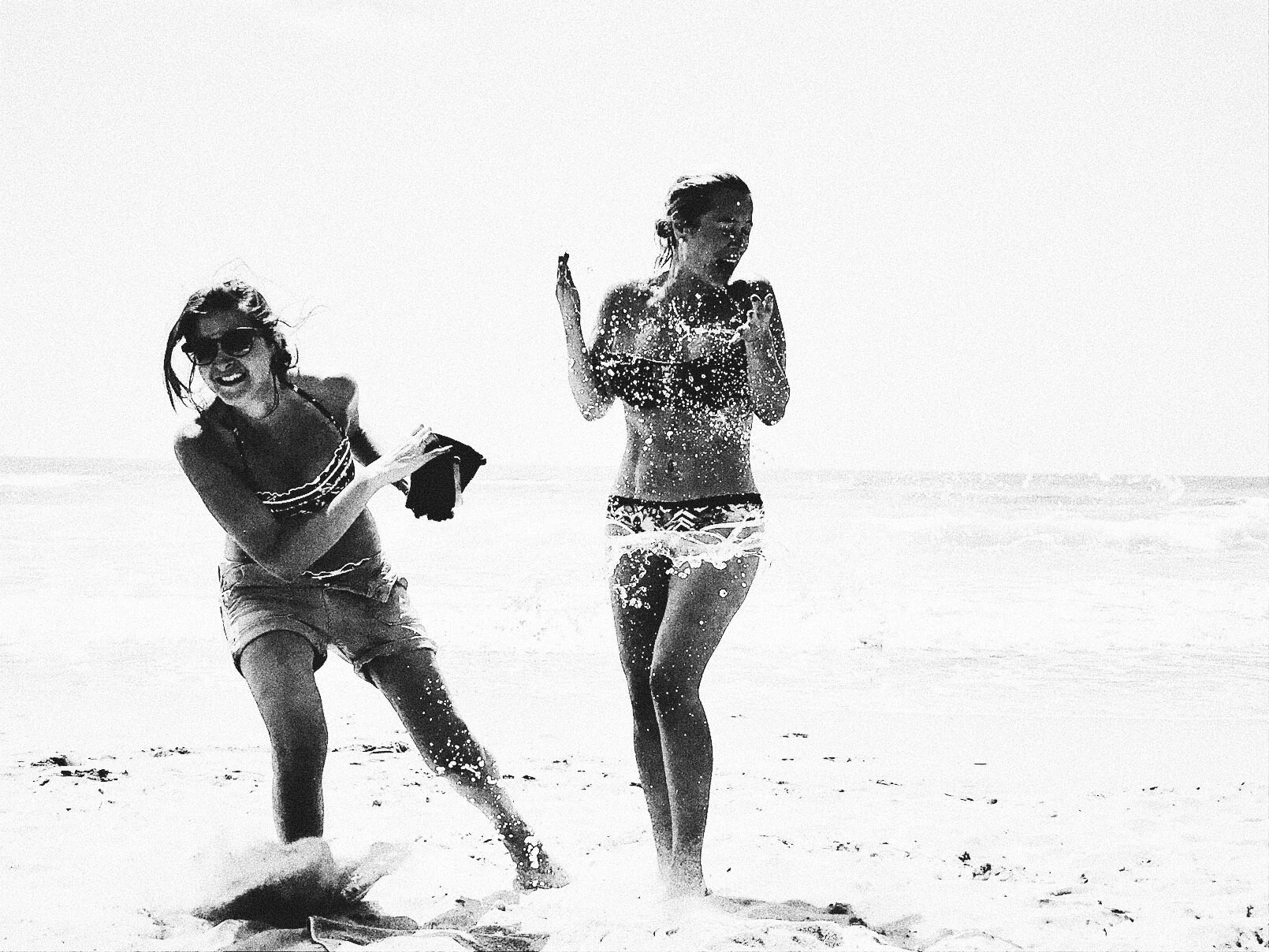 Two women playing on a beach, splashing water and laughing, with the ocean in the background, in a black and white photo.