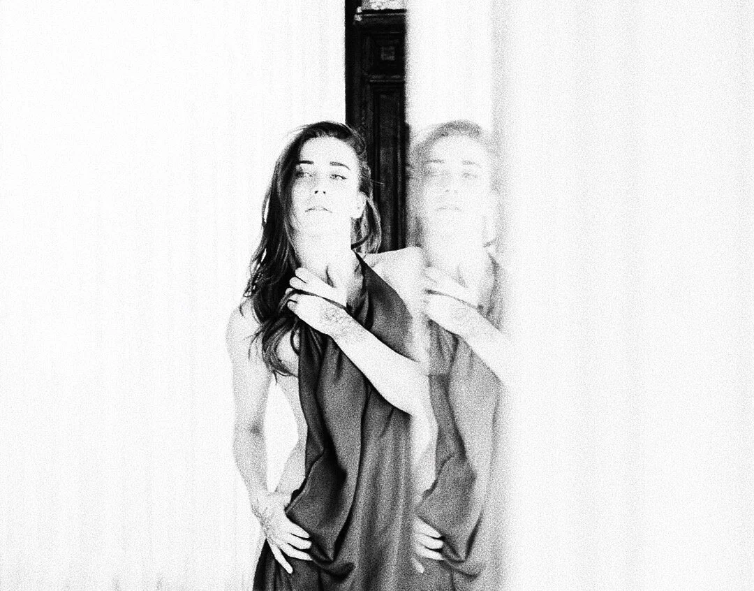 Black and white blurred image of a person with long hair, holding or adjusting a garment, standing near a doorway. The image has a mirror effect with a double exposure.