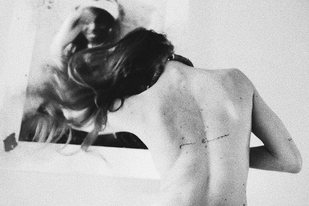 Black and white photo of a person with long hair, viewed from behind, with a tattoo on their upper back, facing an abstract artwork on the wall.